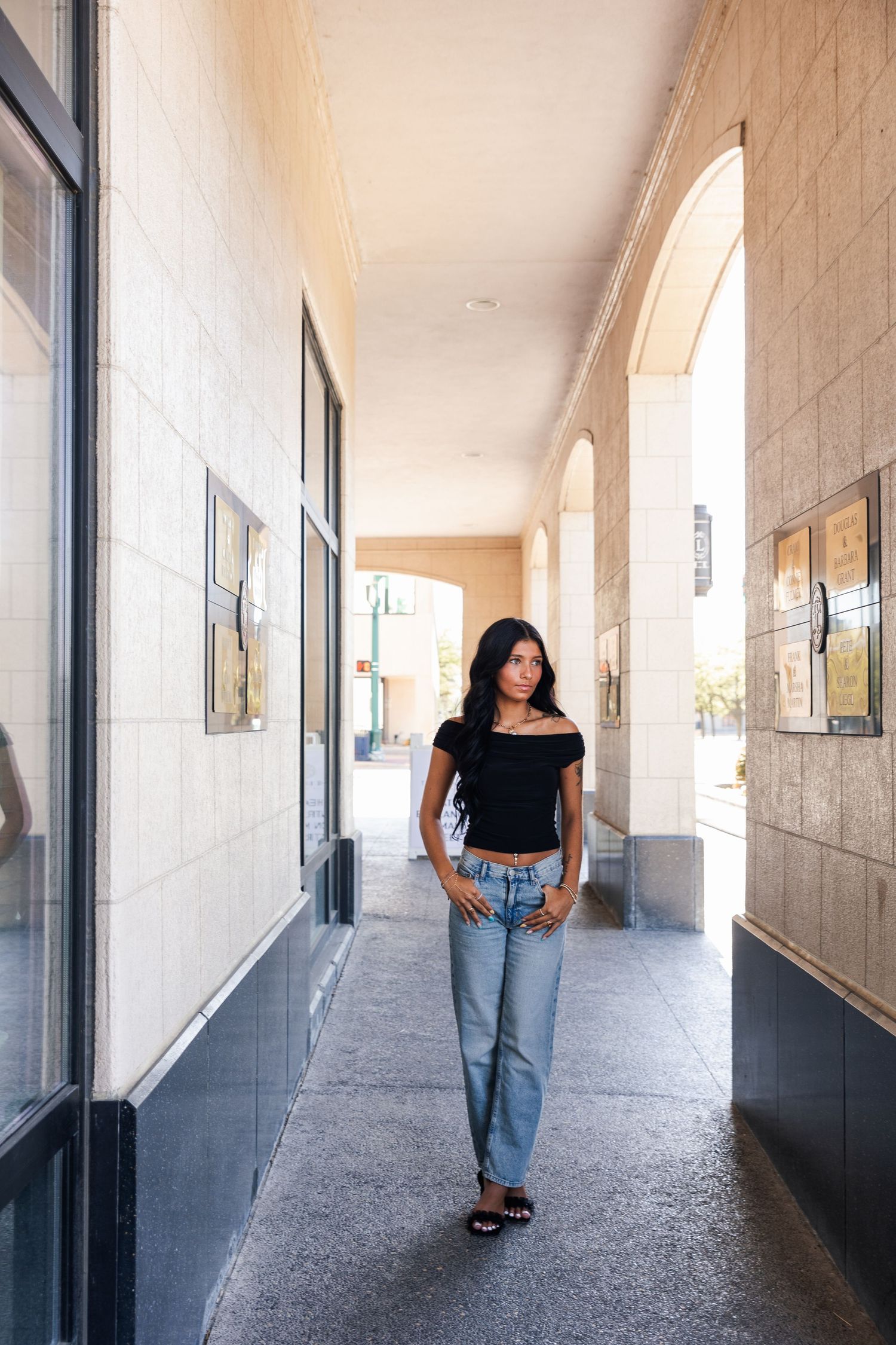 Young woman in black off-shoulder crop top and jeans posing in tiled building corridor.