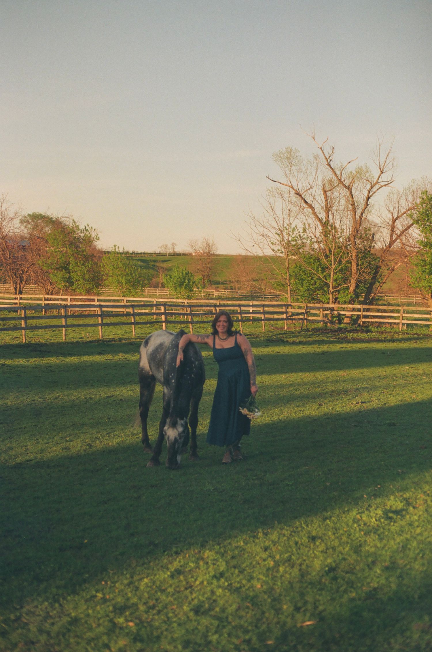 Woman in teal dress walking with a gray horse across a sunlit fenced pasture.
