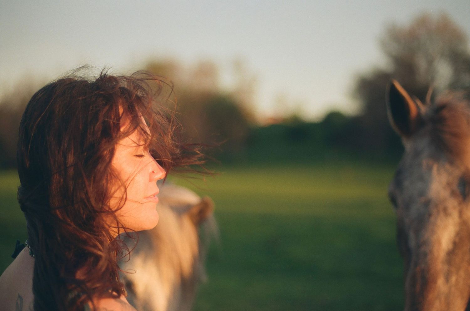 Redhead woman with closed eyes and wind-blown hair near a horse in a sunlit green field.