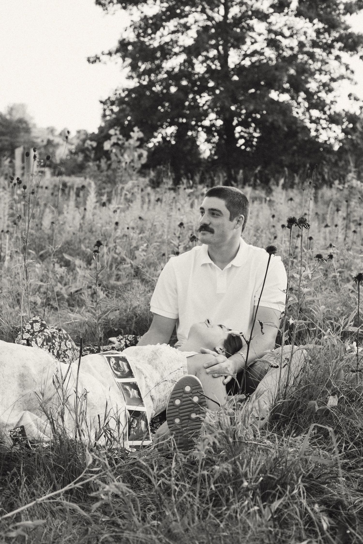 Black and white photo of expectant father sitting in meadow holding ultrasound photos during maternity session.