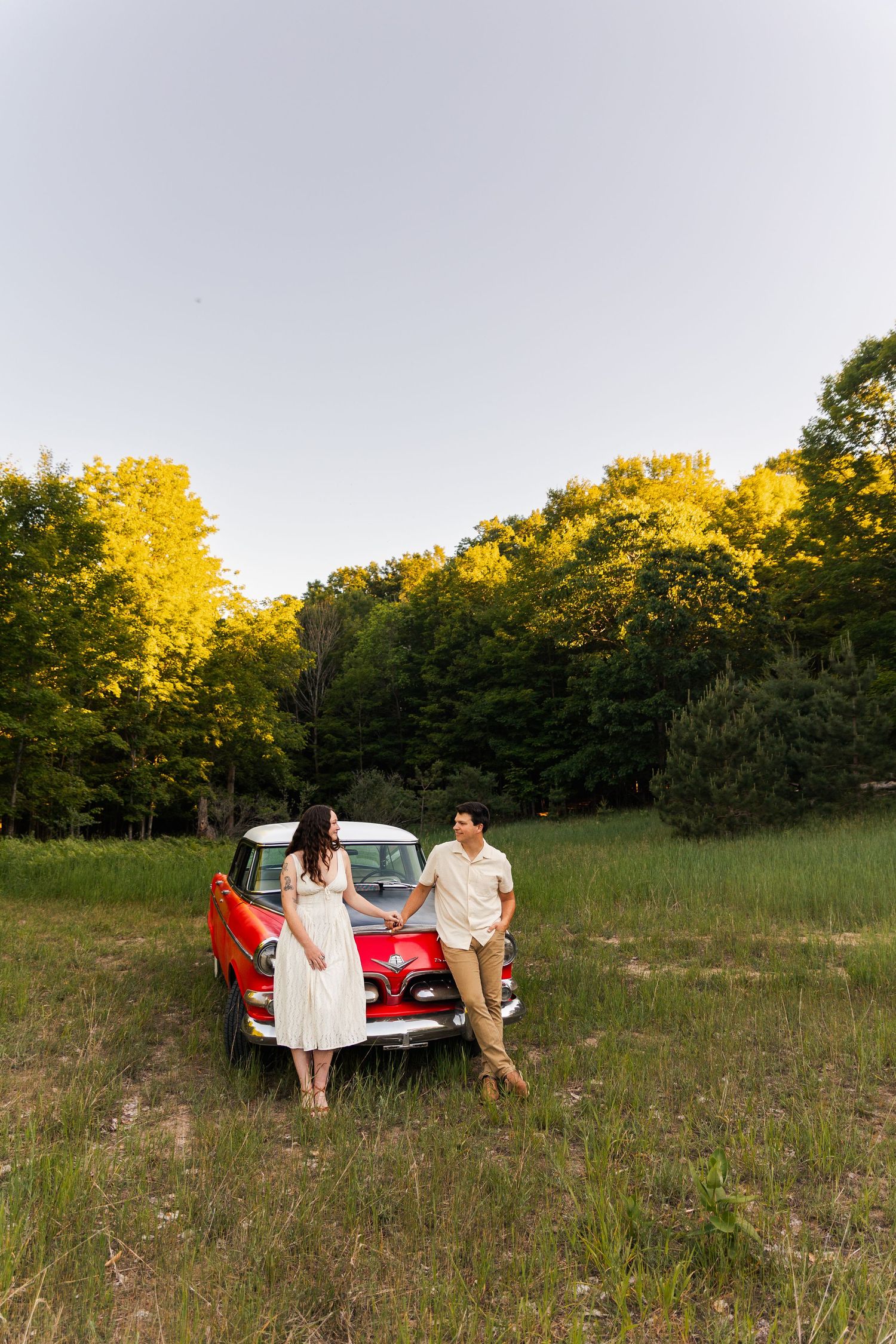 A couple stands together beside a classic red vintage car in a forest clearing at dusk.