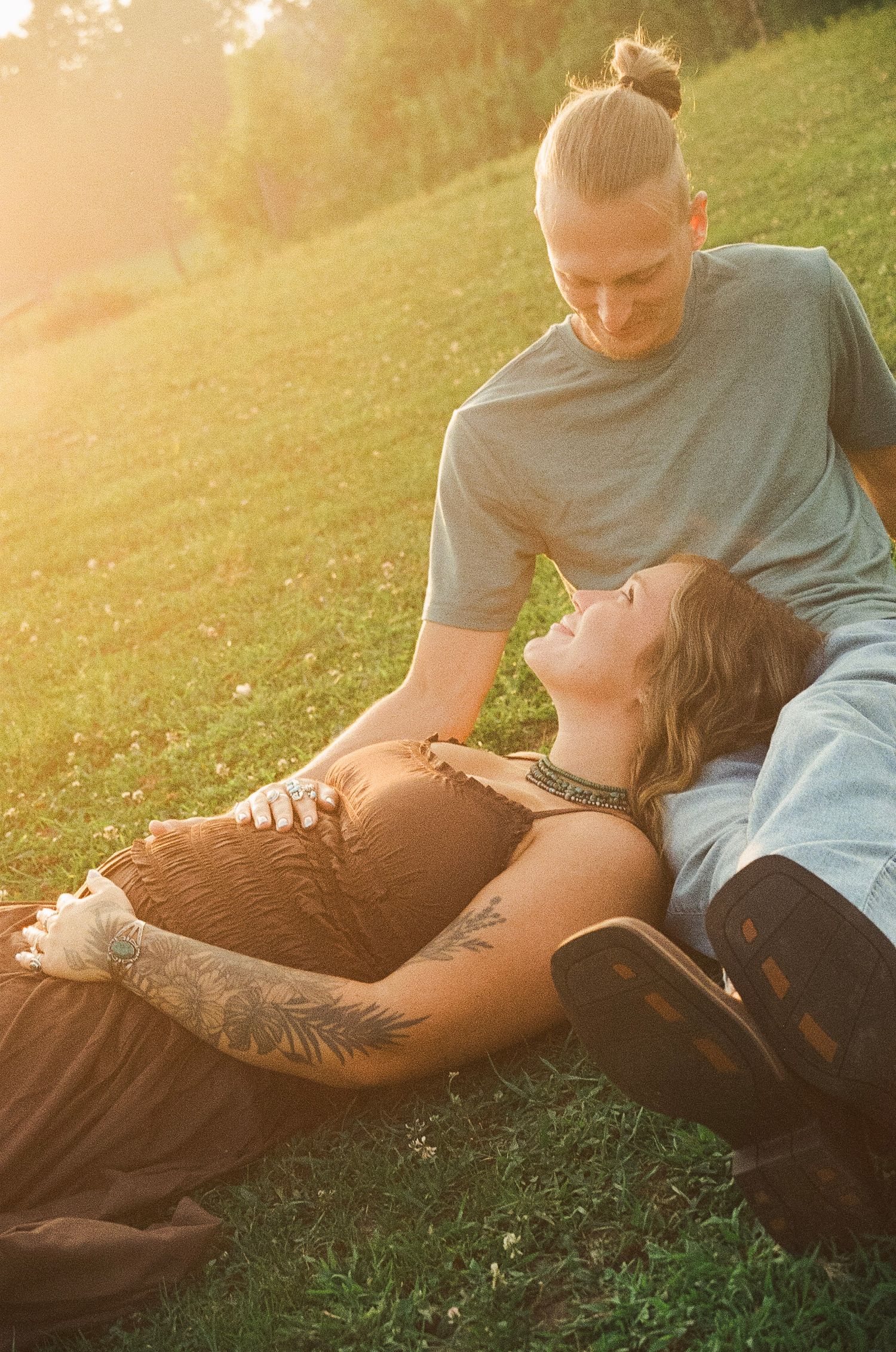 Pregnant woman lying on grass in golden sunlight, resting her head on a man's lap while he gazes at her baby bump.