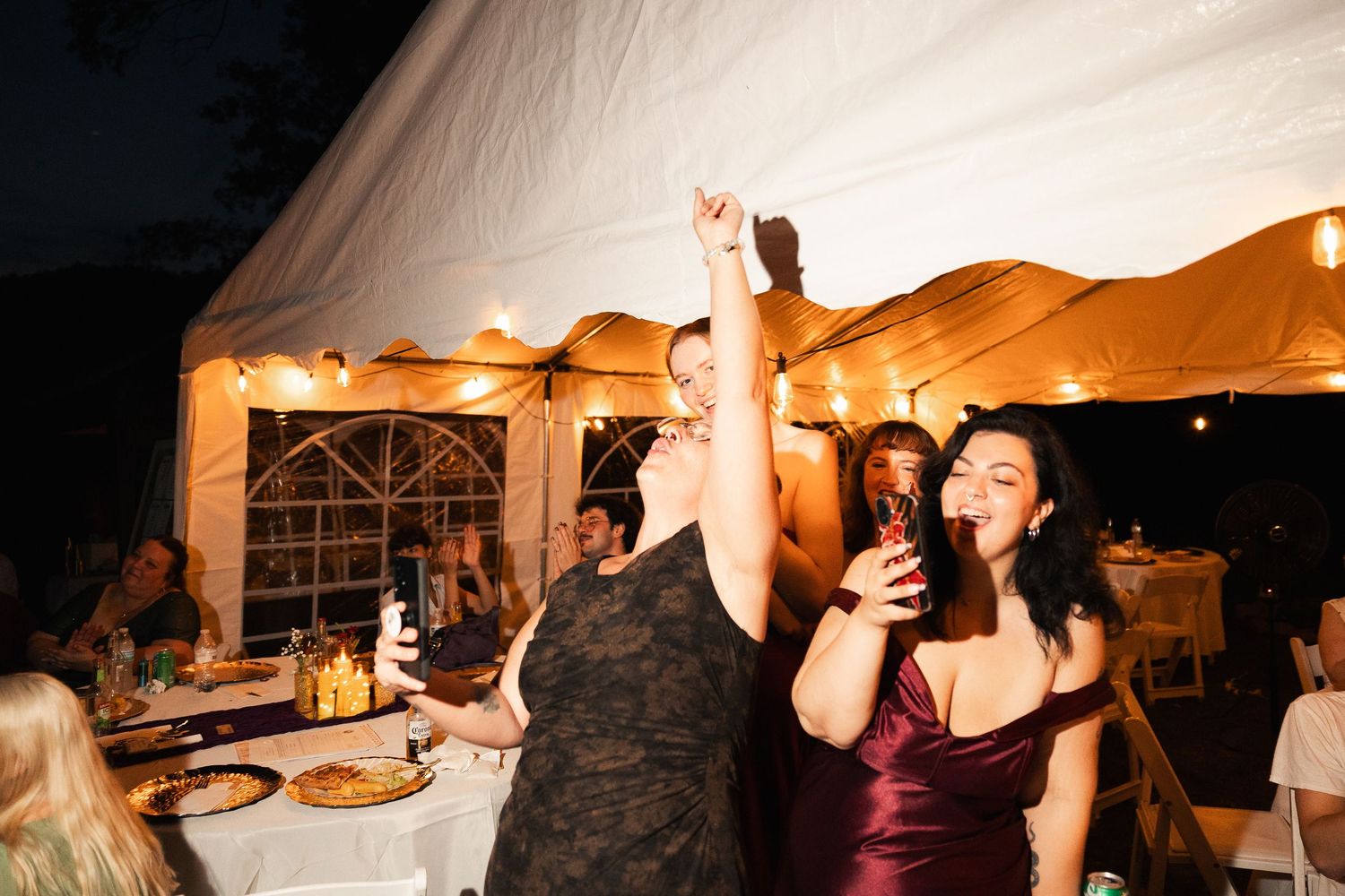 Women dancing and celebrating at an outdoor evening wedding reception under a white tent with string lights.