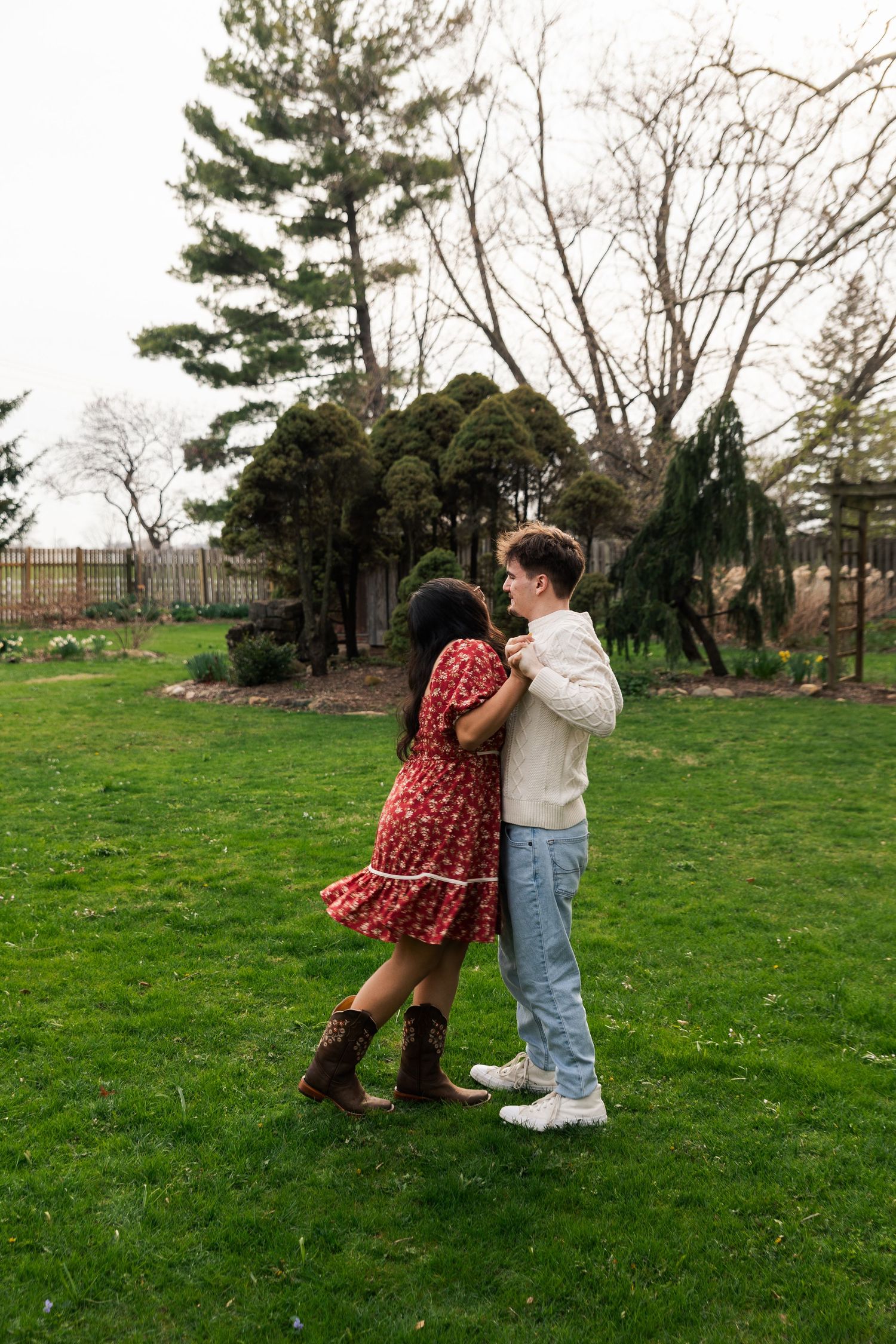 Young couple dancing and embracing on a green lawn in a garden with tall trees.