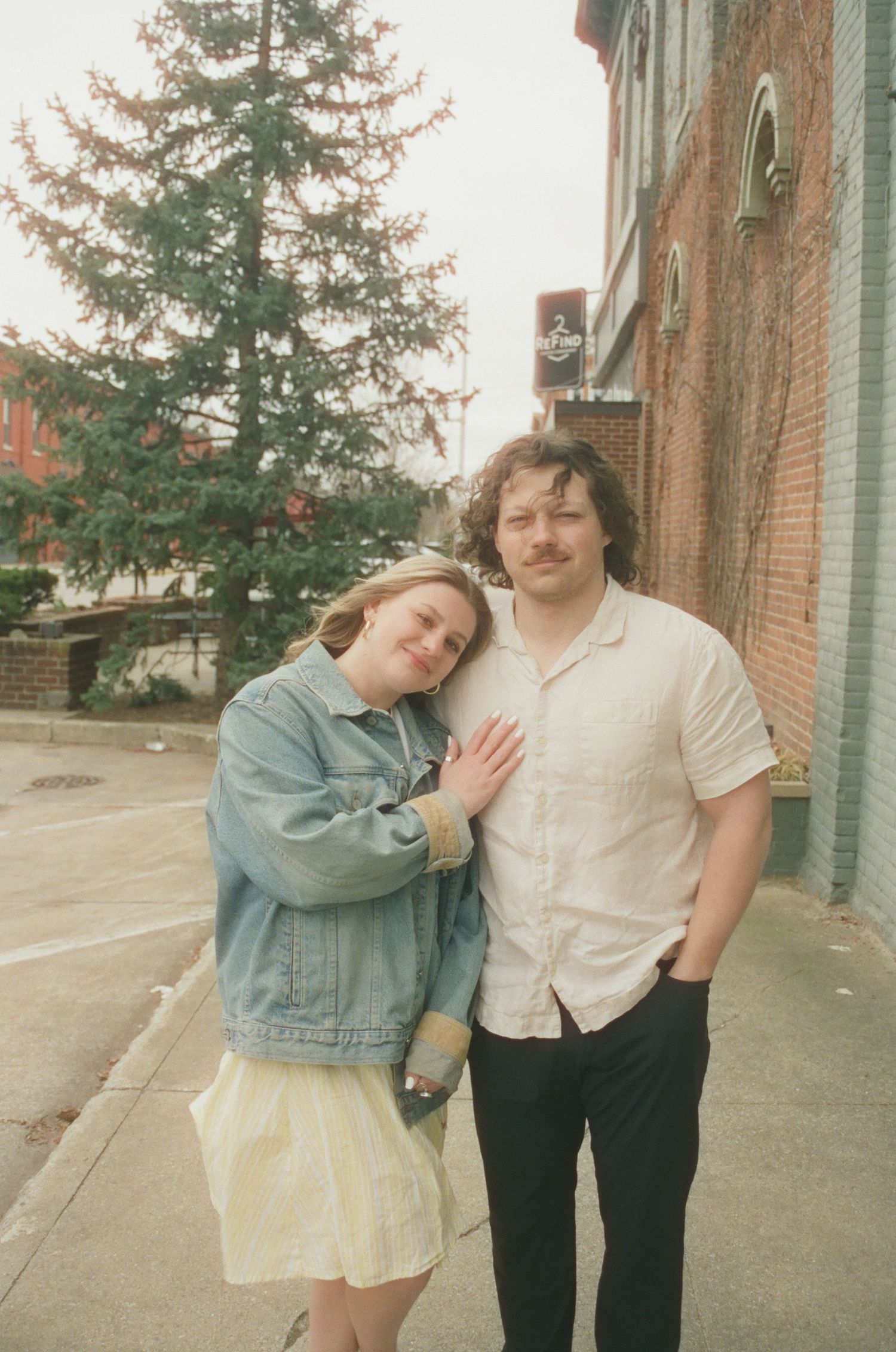 Smiling couple posing on urban sidewalk, woman in denim jacket leaning on mans shoulder.