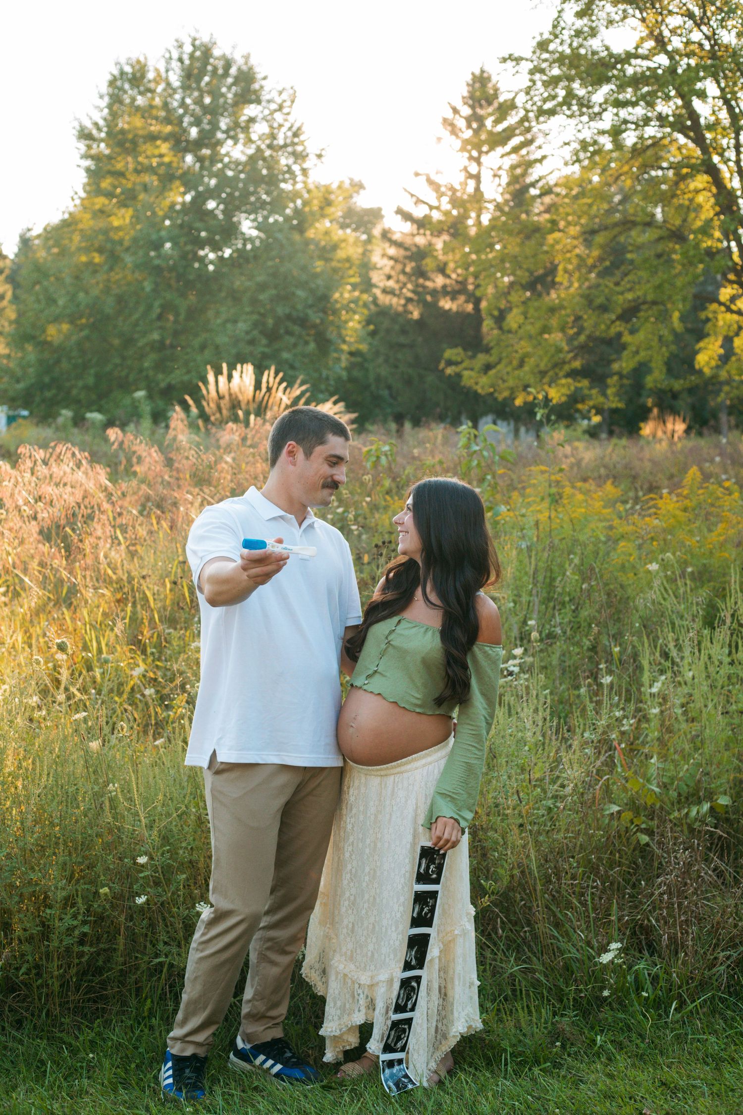 Couple holding ultrasound photos in sunlit field during outdoor maternity photography session.