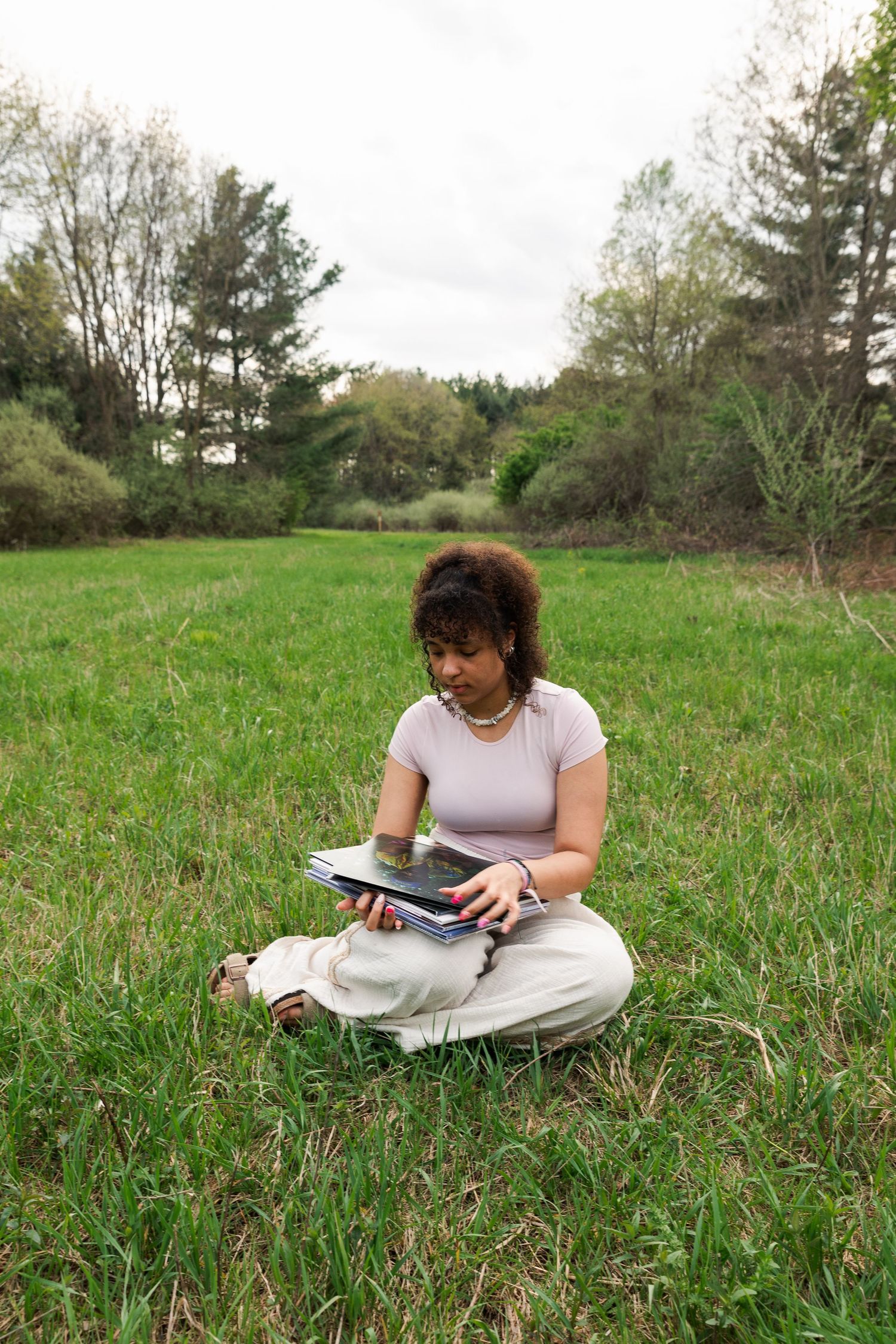 Young woman with curly hair sitting cross-legged on grass in open field, reading books outdoors.