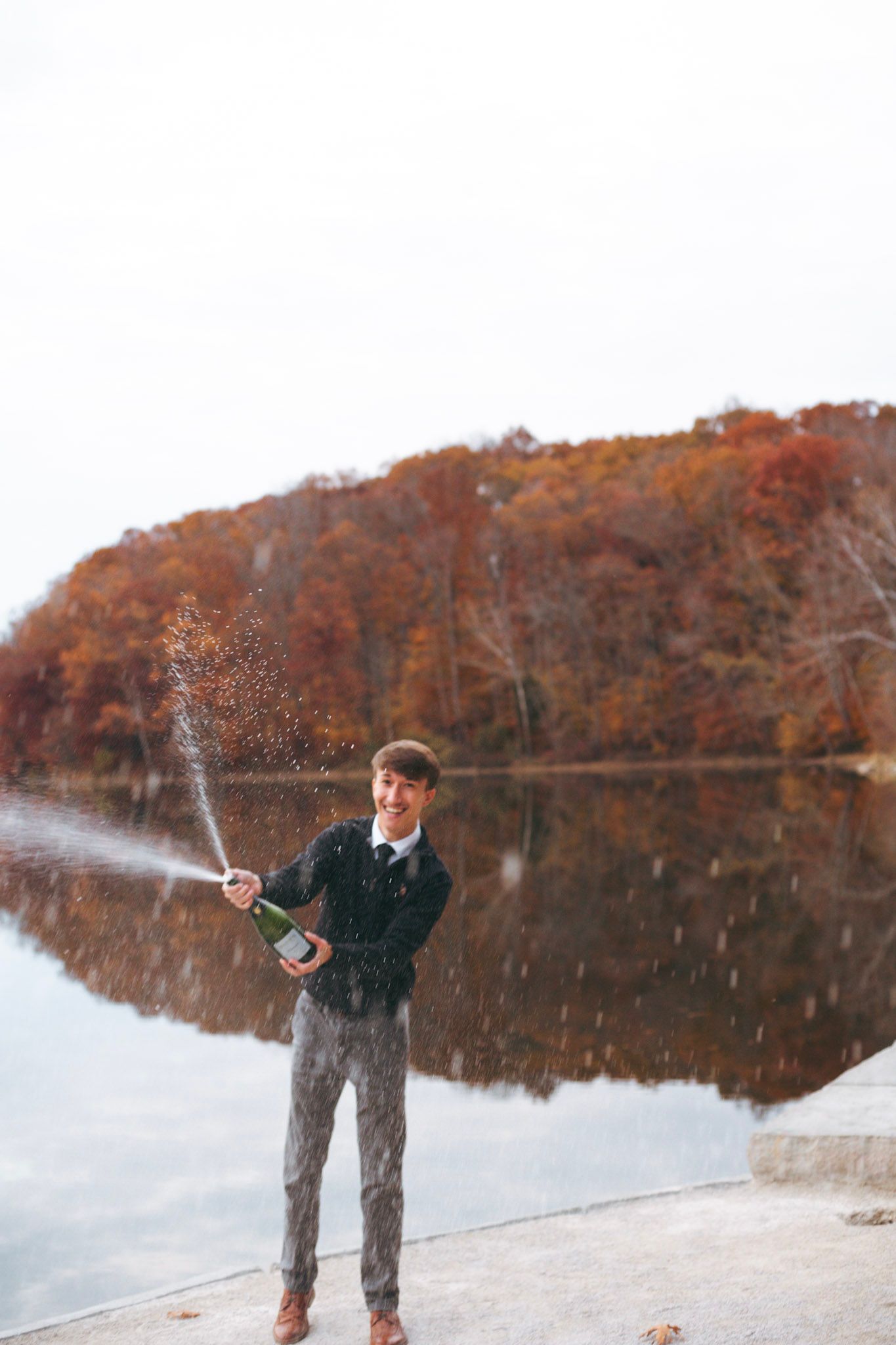 Young man in formal attire popping champagne bottle by a lake with vibrant fall foliage in background.