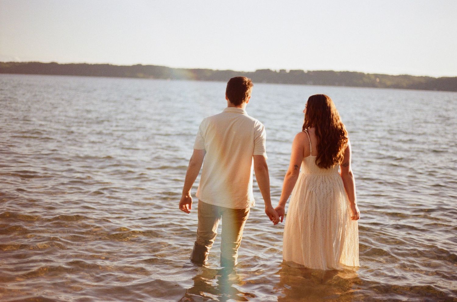 A couple holds hands while wading through shallow water at sunset, viewed from behind.
