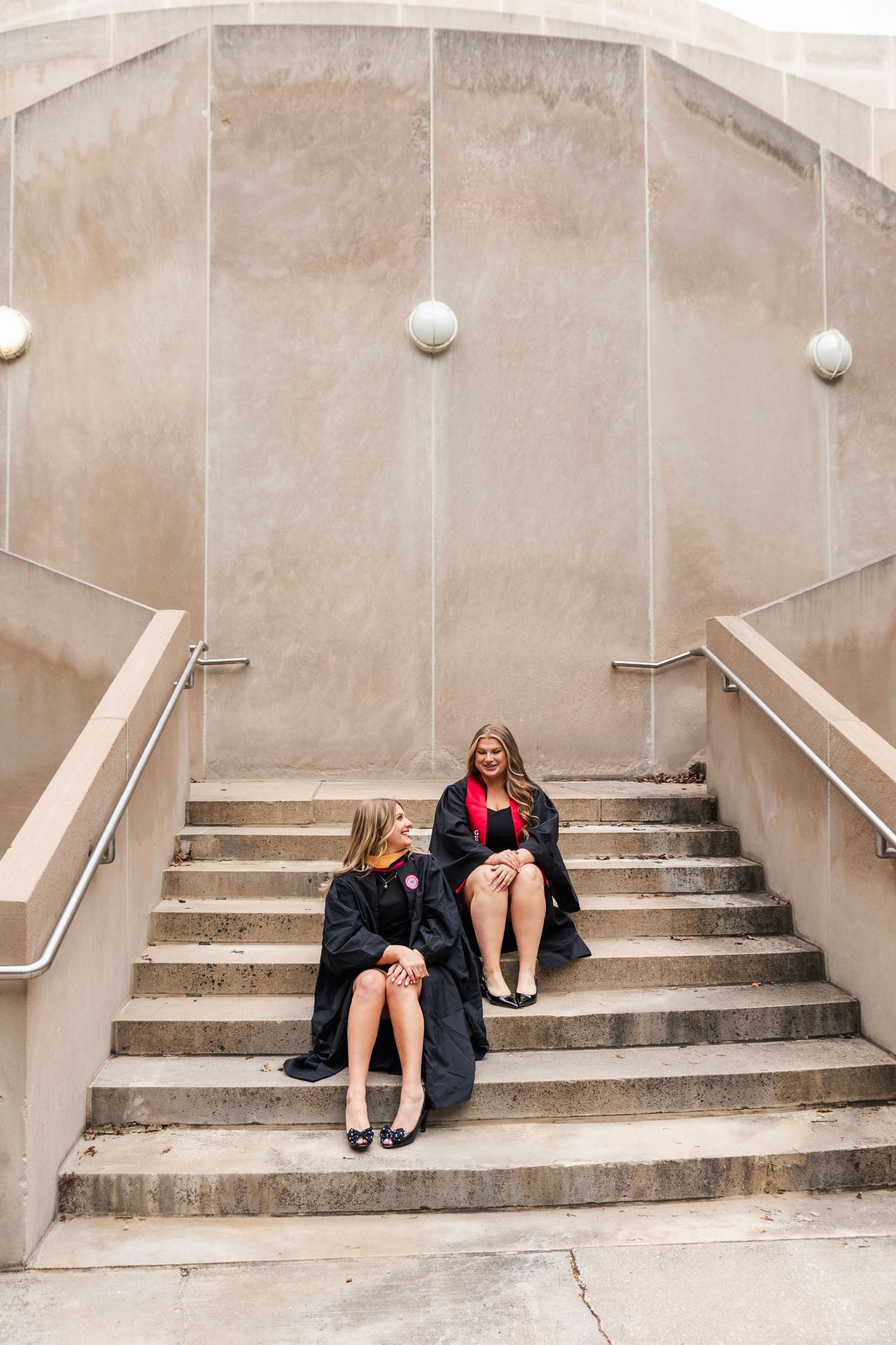 Two graduates in black academic robes sitting and chatting on concrete outdoor staircase steps.