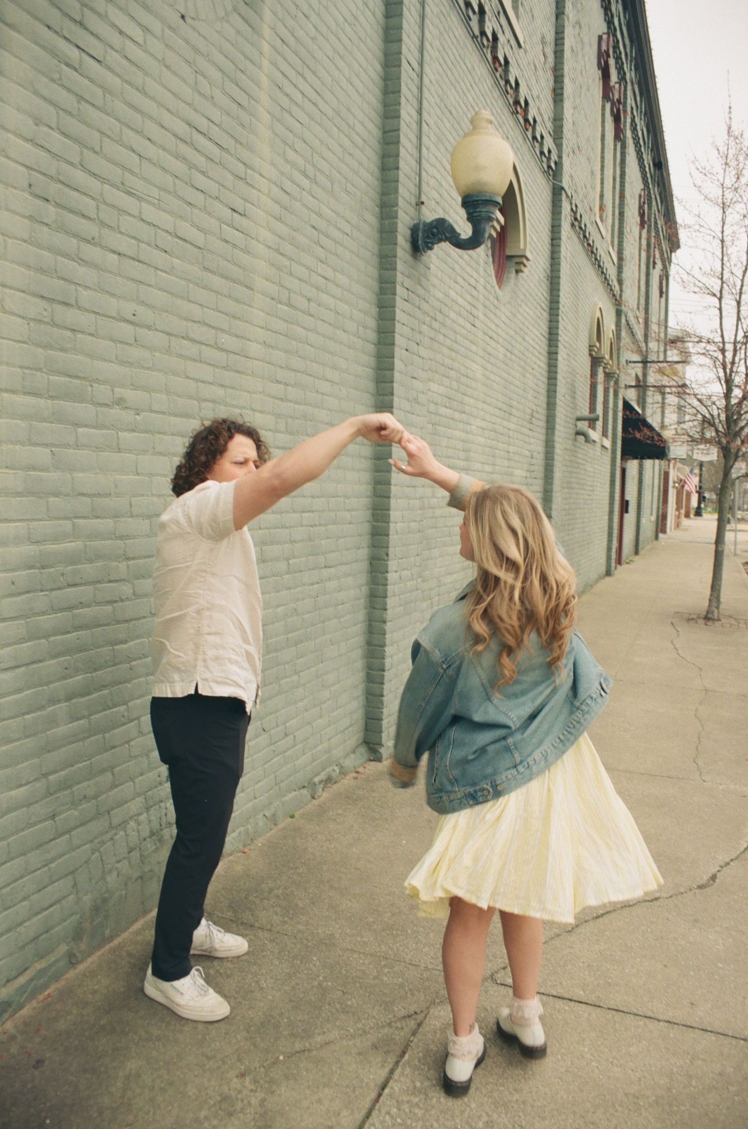 Couple dancing and spinning on a city sidewalk, woman in yellow skirt and denim jacket twirling.