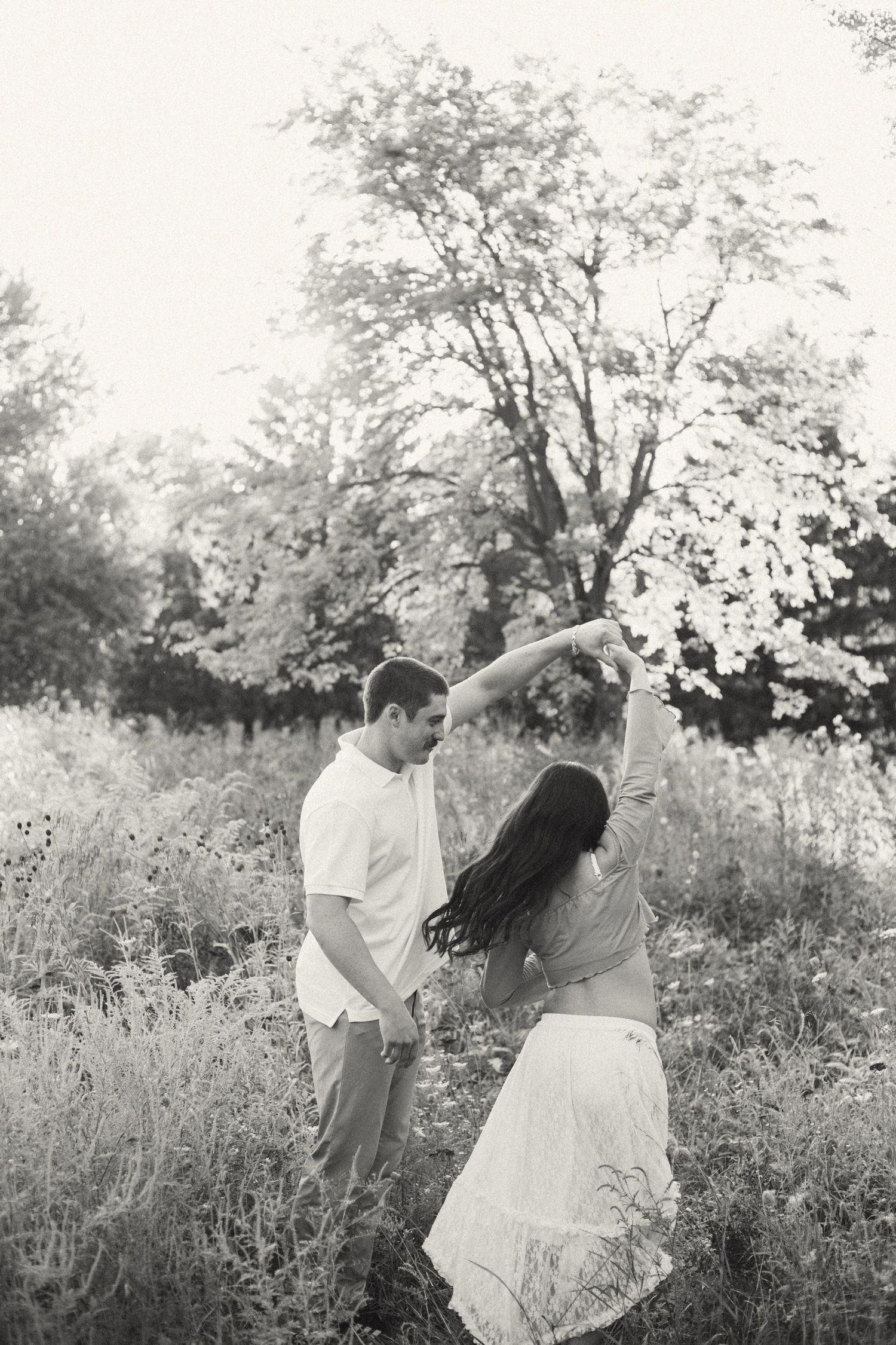 Black and white photo of expectant couple dancing together in a meadow surrounded by tall trees.