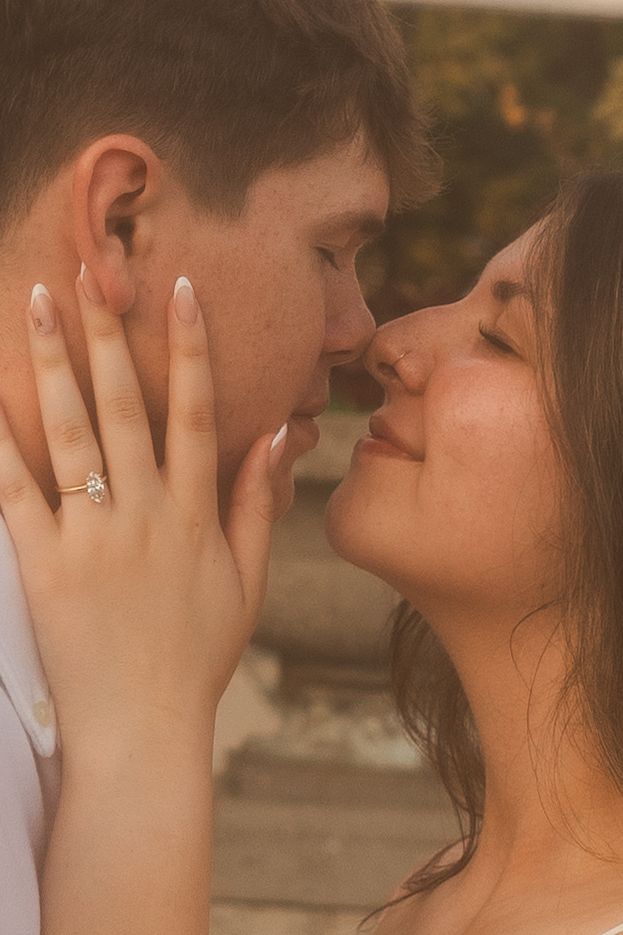 Couple sharing a romantic almost-kiss moment, woman wearing diamond engagement ring, warm golden toned photo.