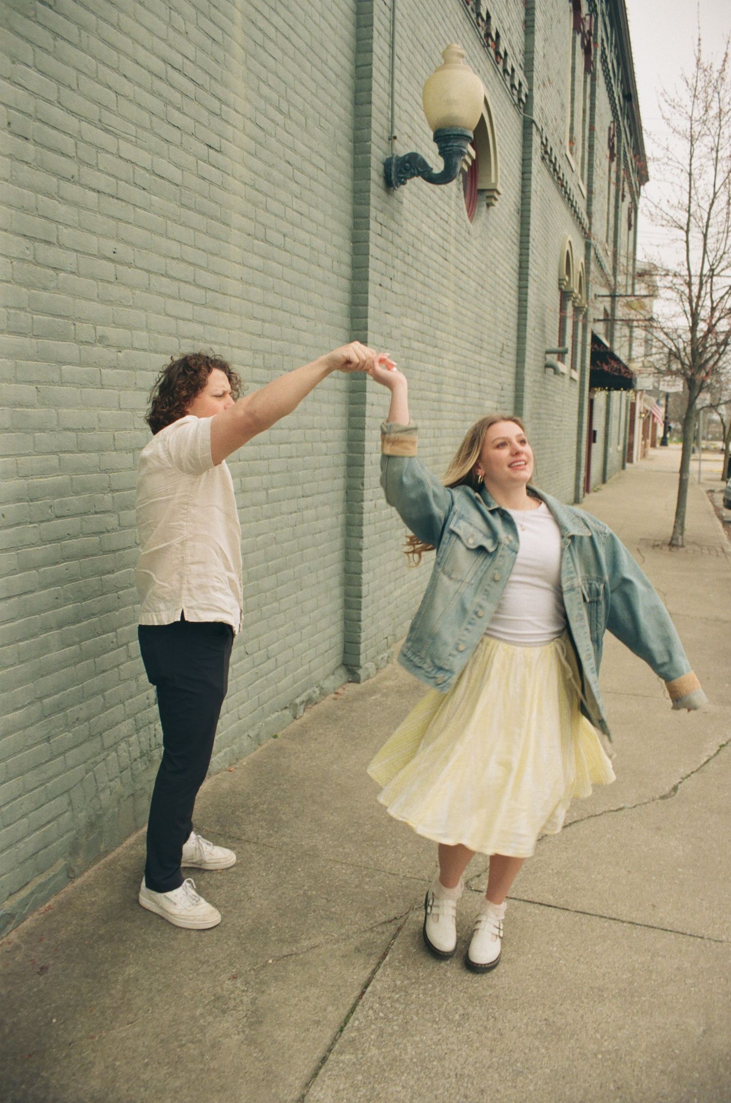 Joyful couple twirling together on city sidewalk, woman in yellow skirt and denim jacket laughing.