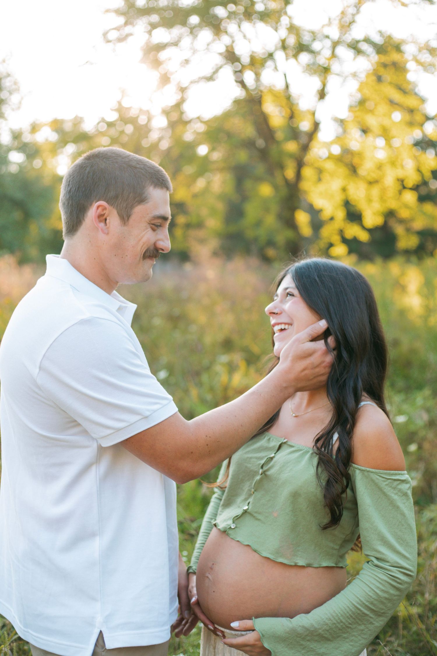 Loving couple gazing at each other as man tenderly touches pregnant woman's face in sunny meadow.