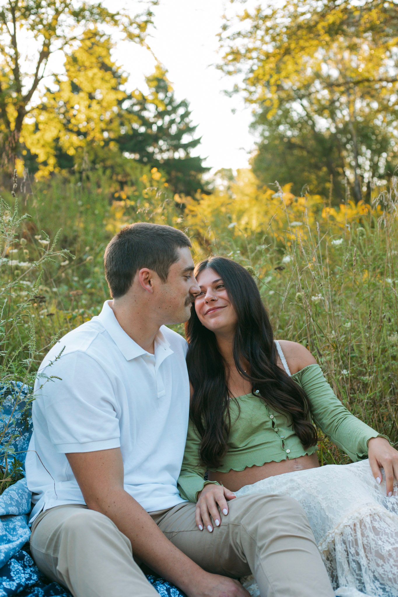 Couple sits together in a golden wildflower meadow, sharing a tender romantic moment at sunset.