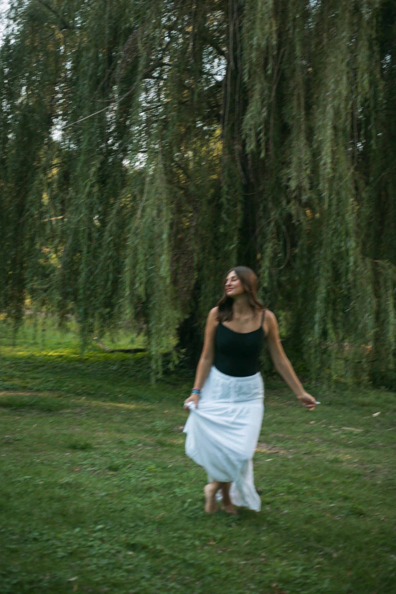 Young woman in black top and white skirt walking barefoot under a weeping willow tree on green grass.
