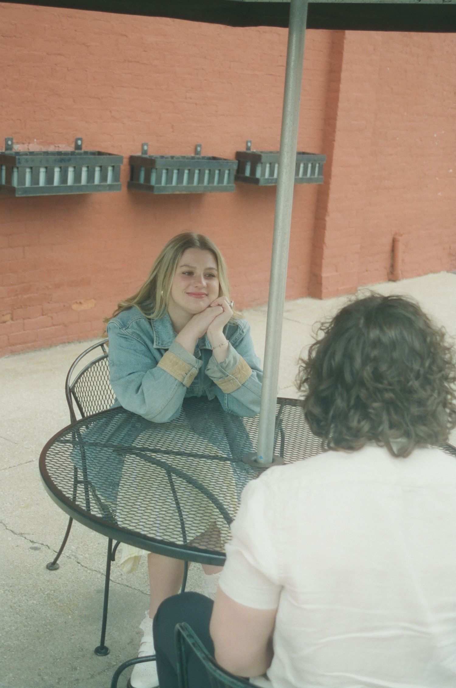 Smiling blonde woman in denim jacket sitting at iron patio table with hands clasped under chin.