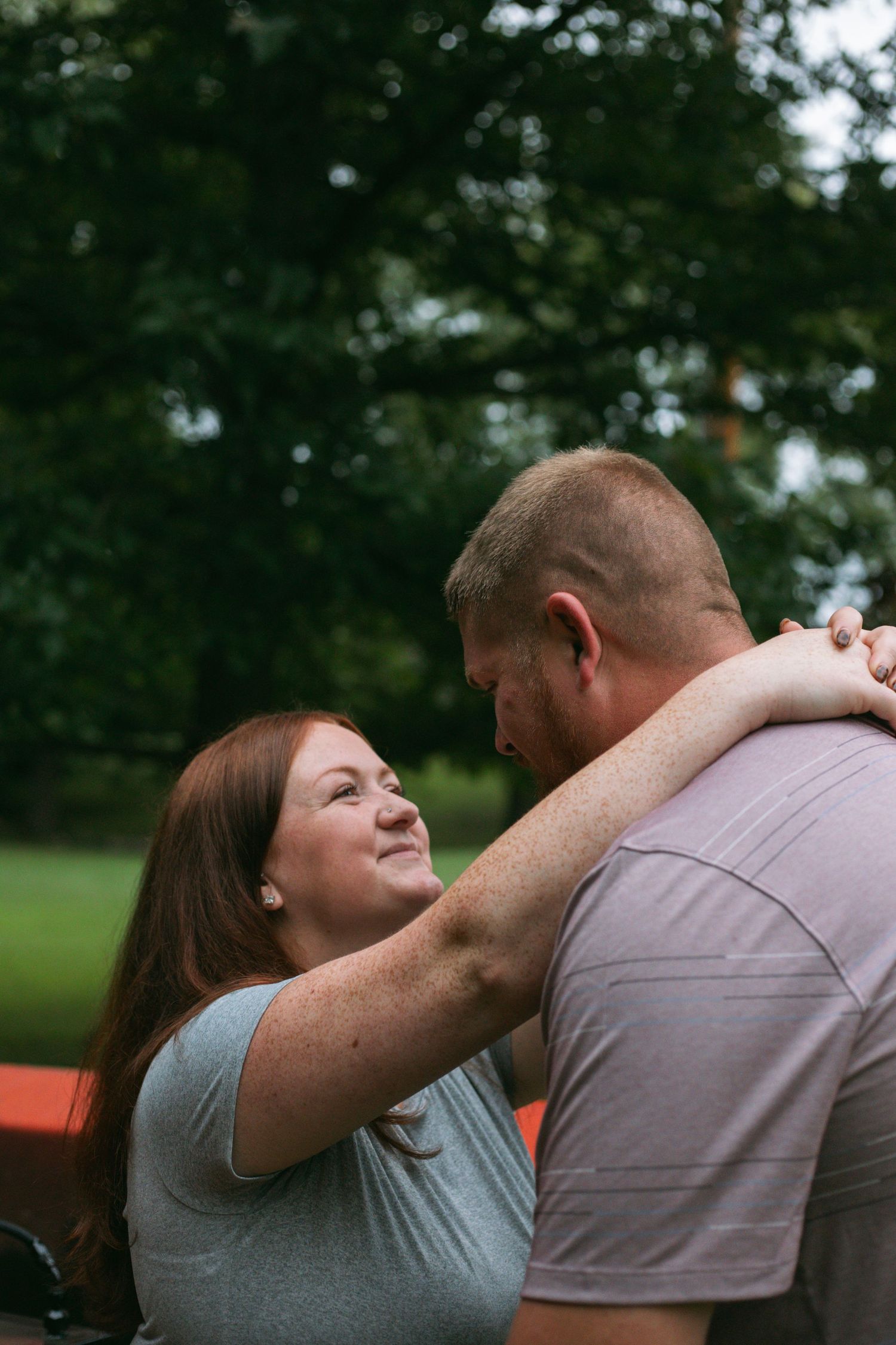 A woman with red hair smiles while embracing a man outdoors with green trees softly blurred in the background.