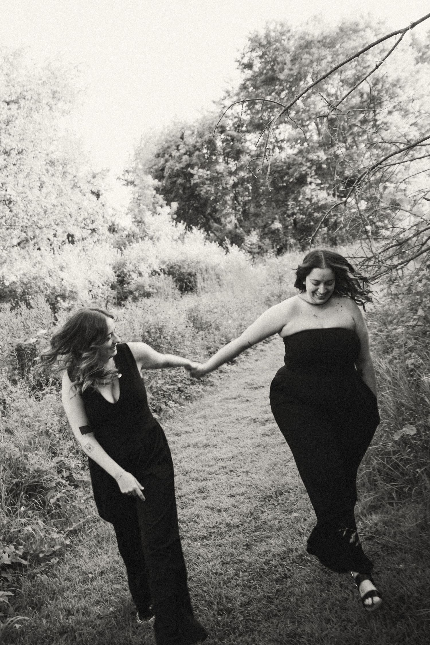 Two women in black outfits hold hands and laugh while walking on a garden path in a black and white photo.
