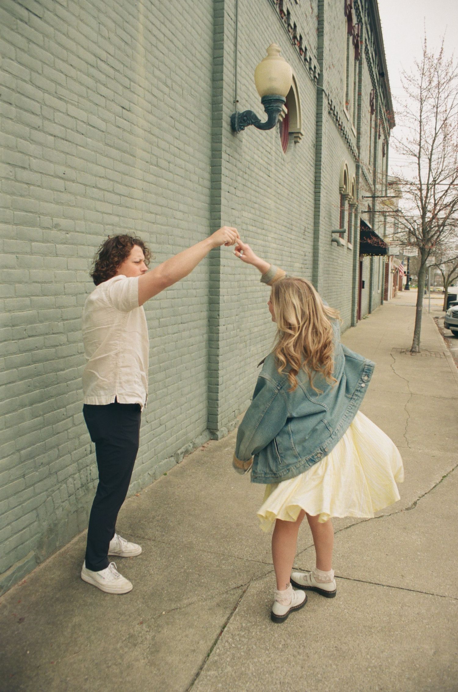 Man spins woman in a yellow dress and denim jacket on a sidewalk beside a green brick wall.