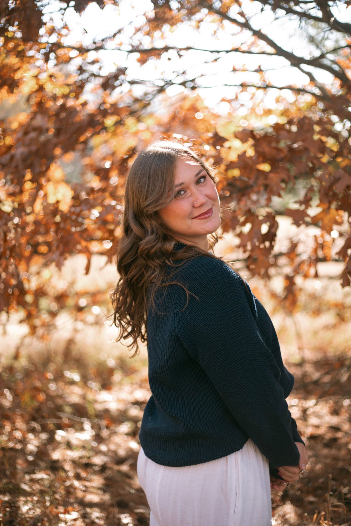 Young woman in navy sweater and white skirt smiling among vibrant autumn foliage in golden light.