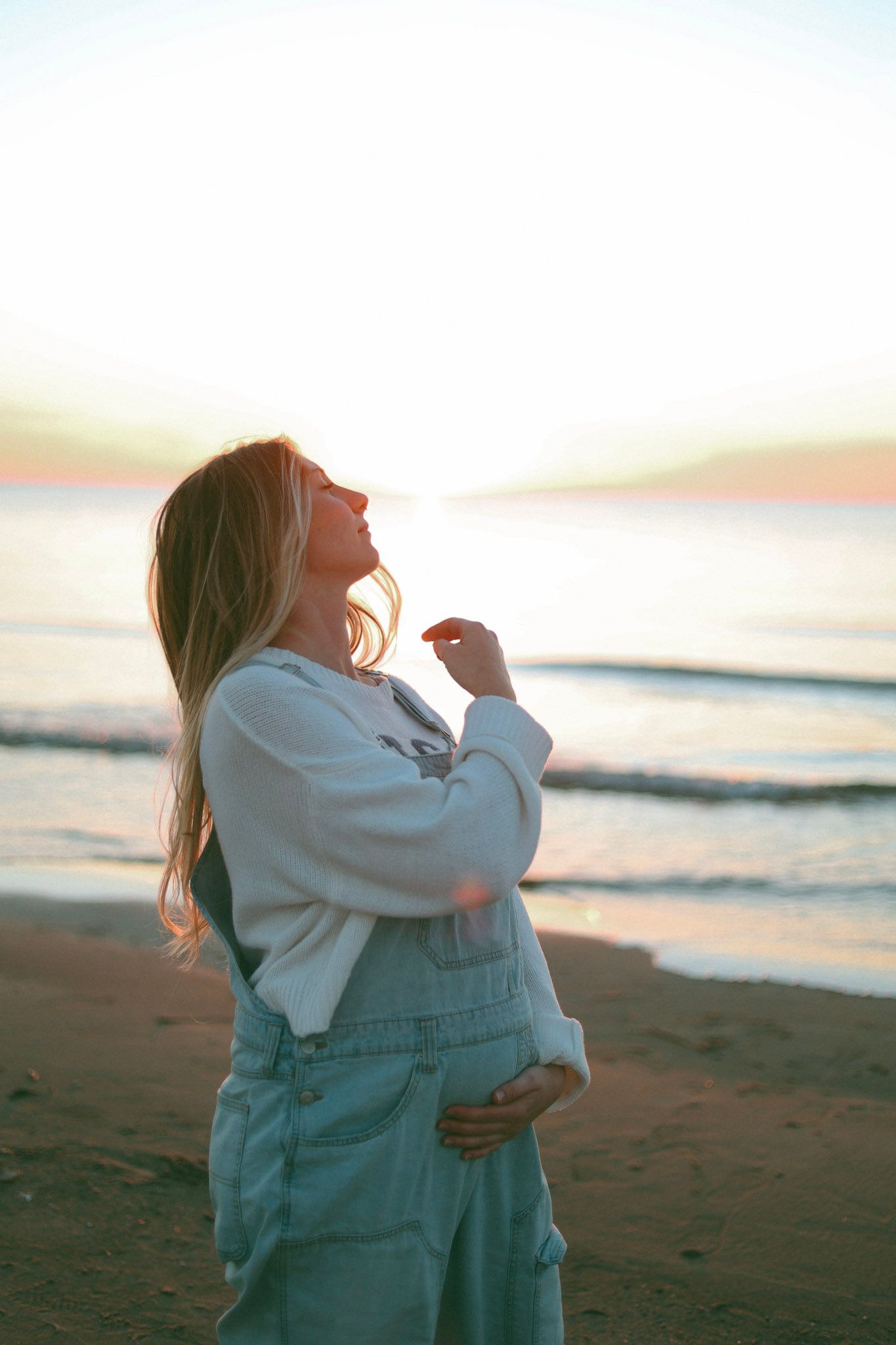 Pregnant woman in denim overalls and white hoodie standing on a beach at golden sunset.
