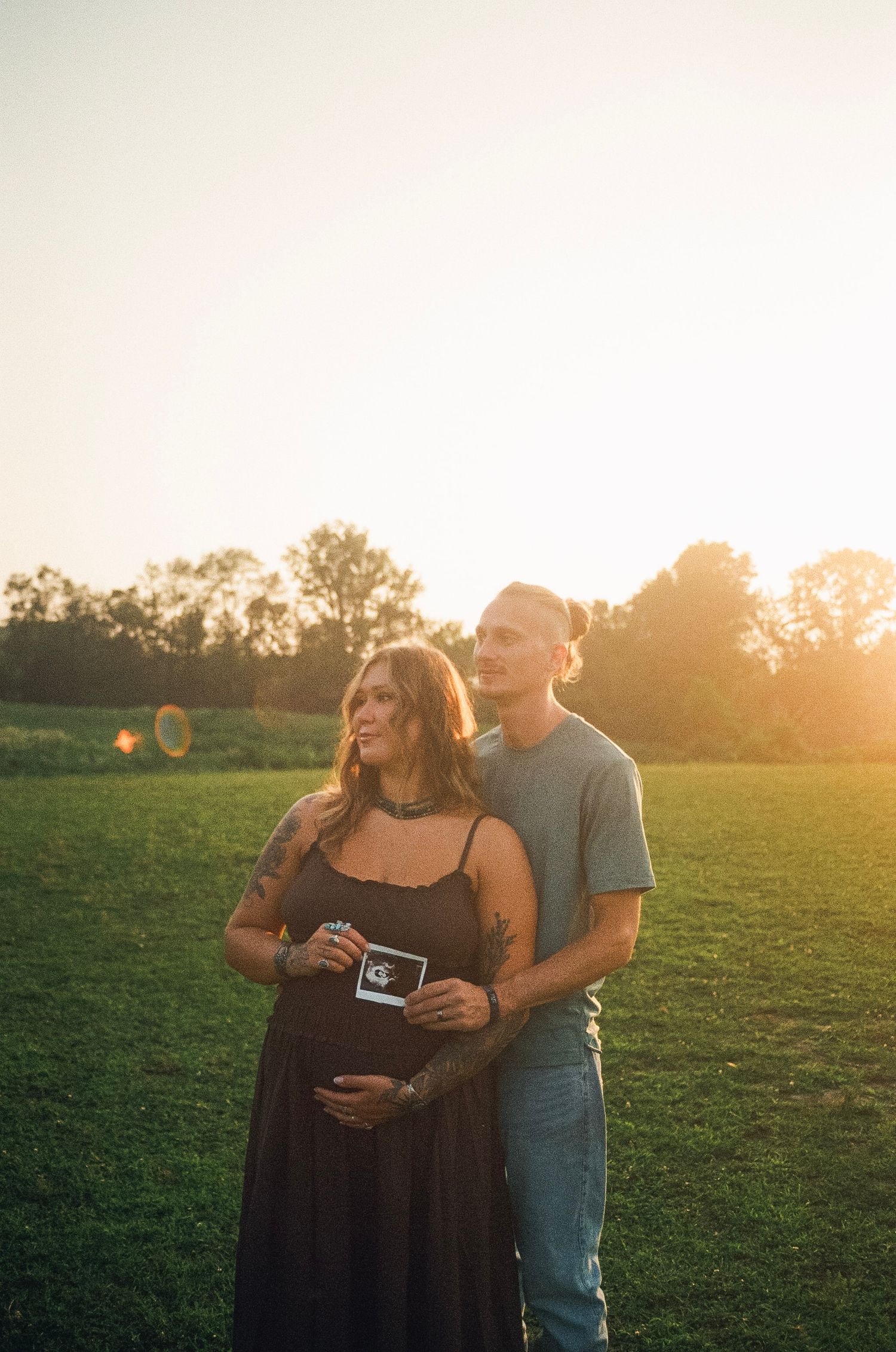 Pregnant couple holding ultrasound photo in golden sunset field, woman in black dress with baby bump, man in gray t-shirt.