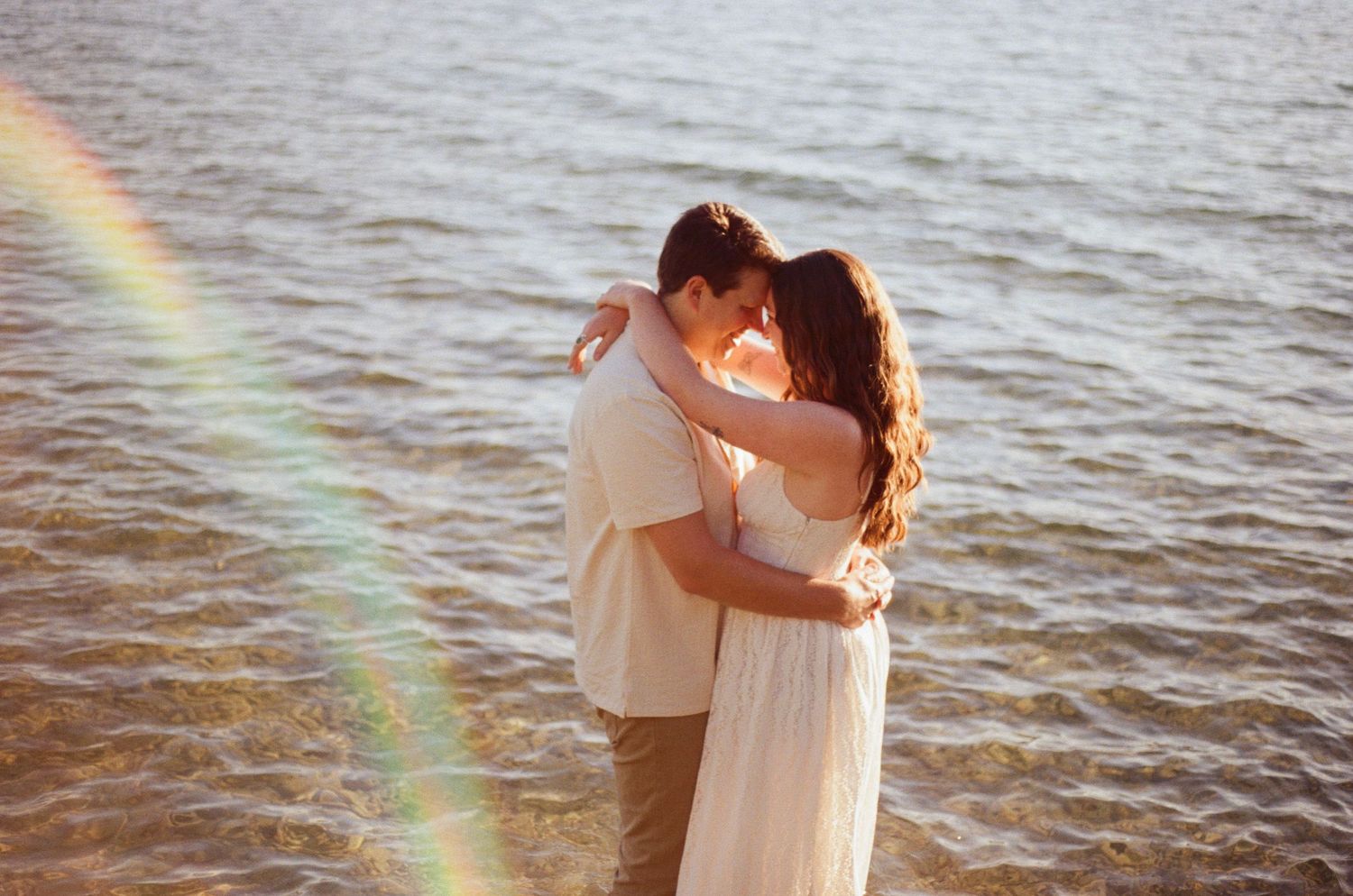 A couple embraces romantically on a sunlit beach with a rainbow lens flare over sparkling water.