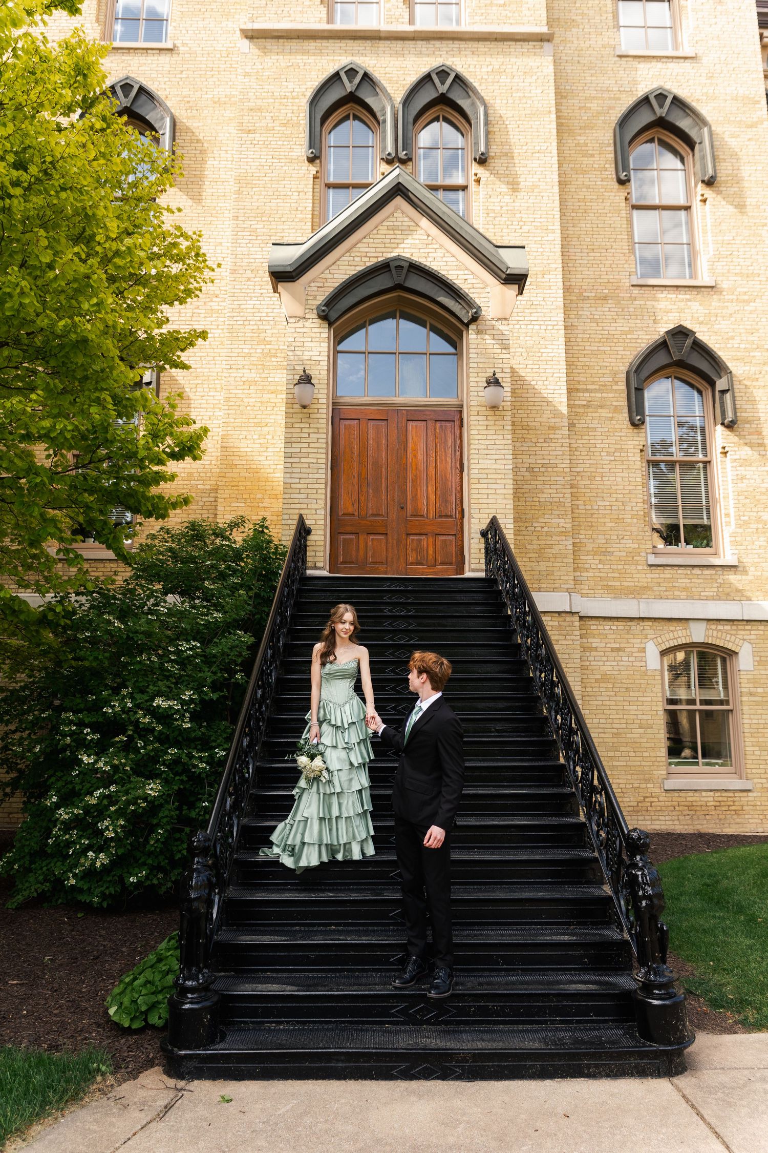 Couple in formal wear posing on black iron staircase outside a Gothic-style yellow brick building.