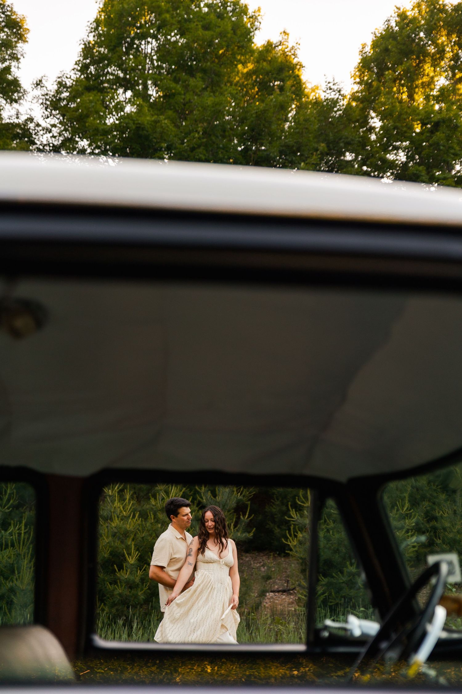 Couple in elegant attire reflected in vintage vehicle mirror, standing in golden meadow with green trees behind.