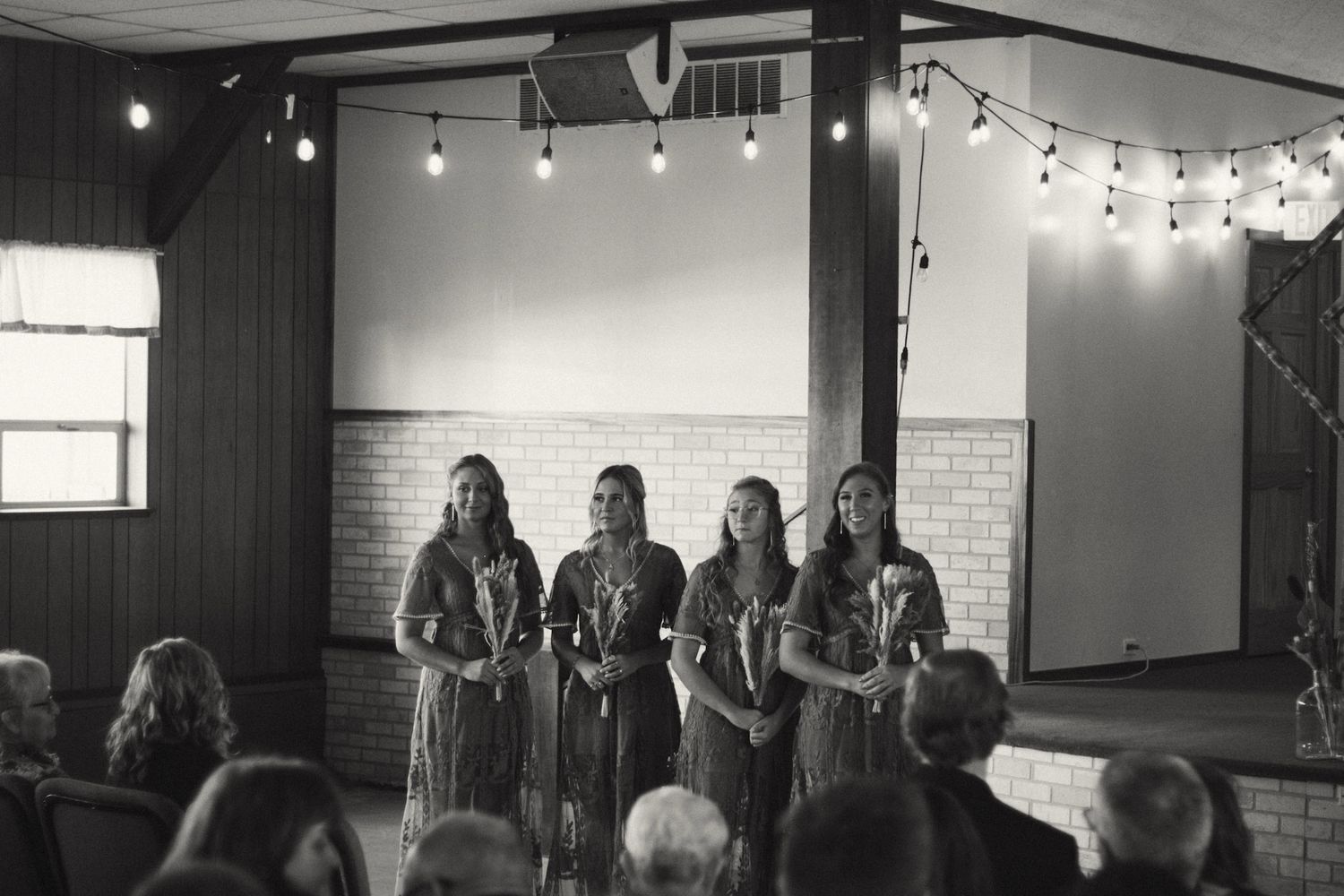 Black and white photo of four bridesmaids standing at wedding ceremony altar holding flowers.