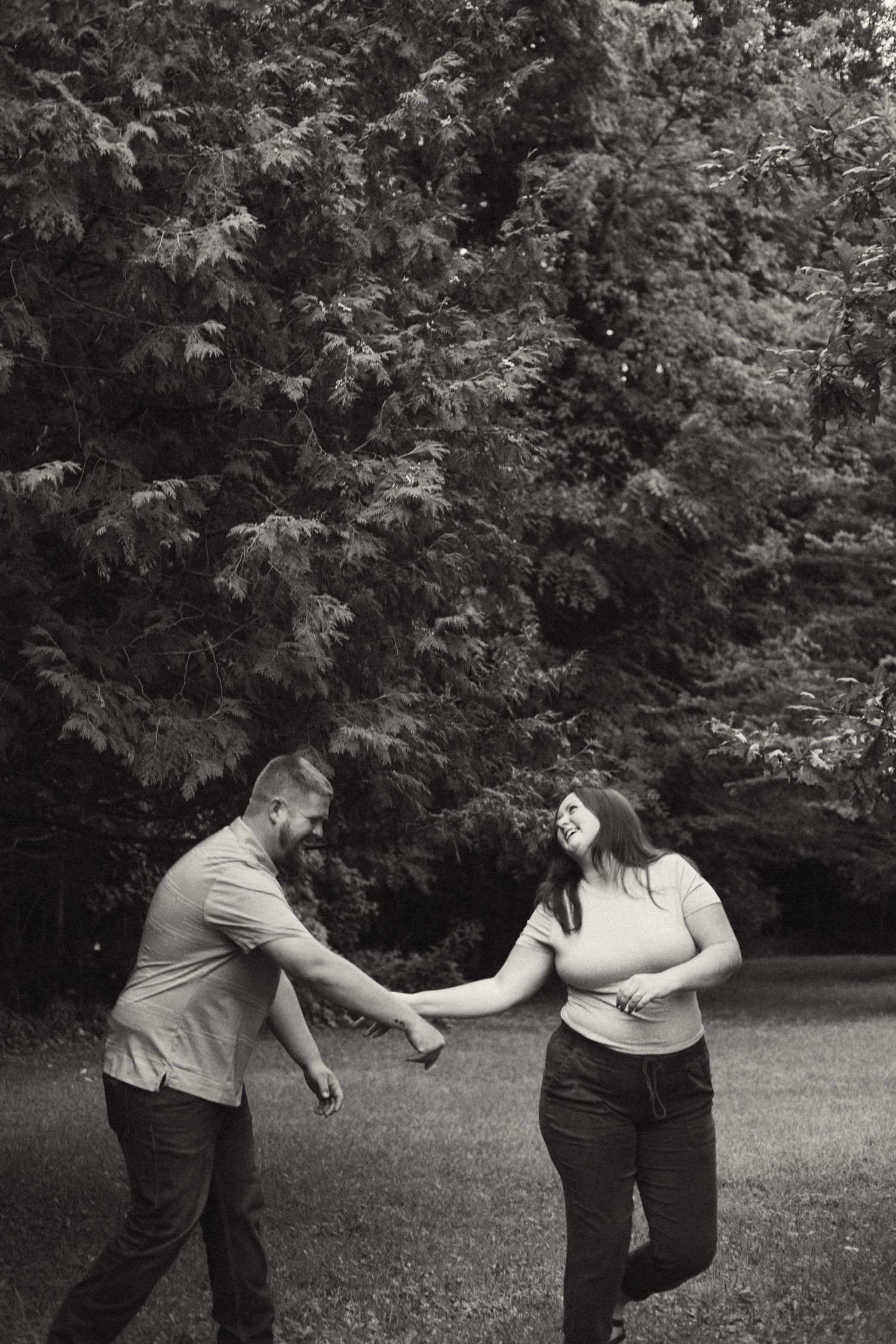 Black and white photo of a laughing couple holding hands and playfully dancing on a grassy lawn.