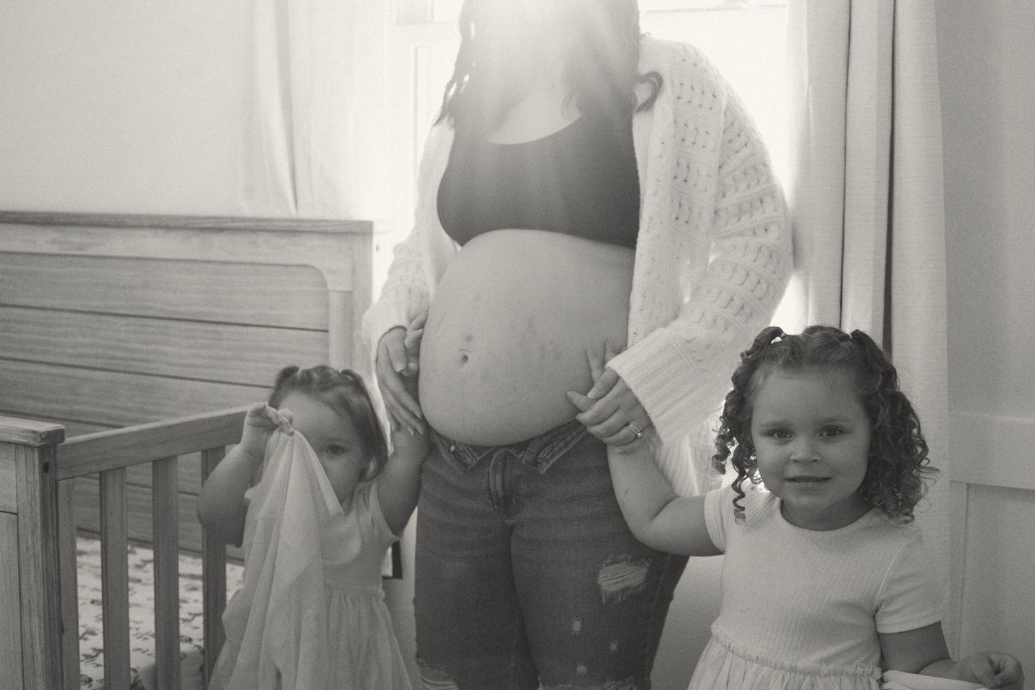 Black and white photo of pregnant woman with two young daughters in nursery room.