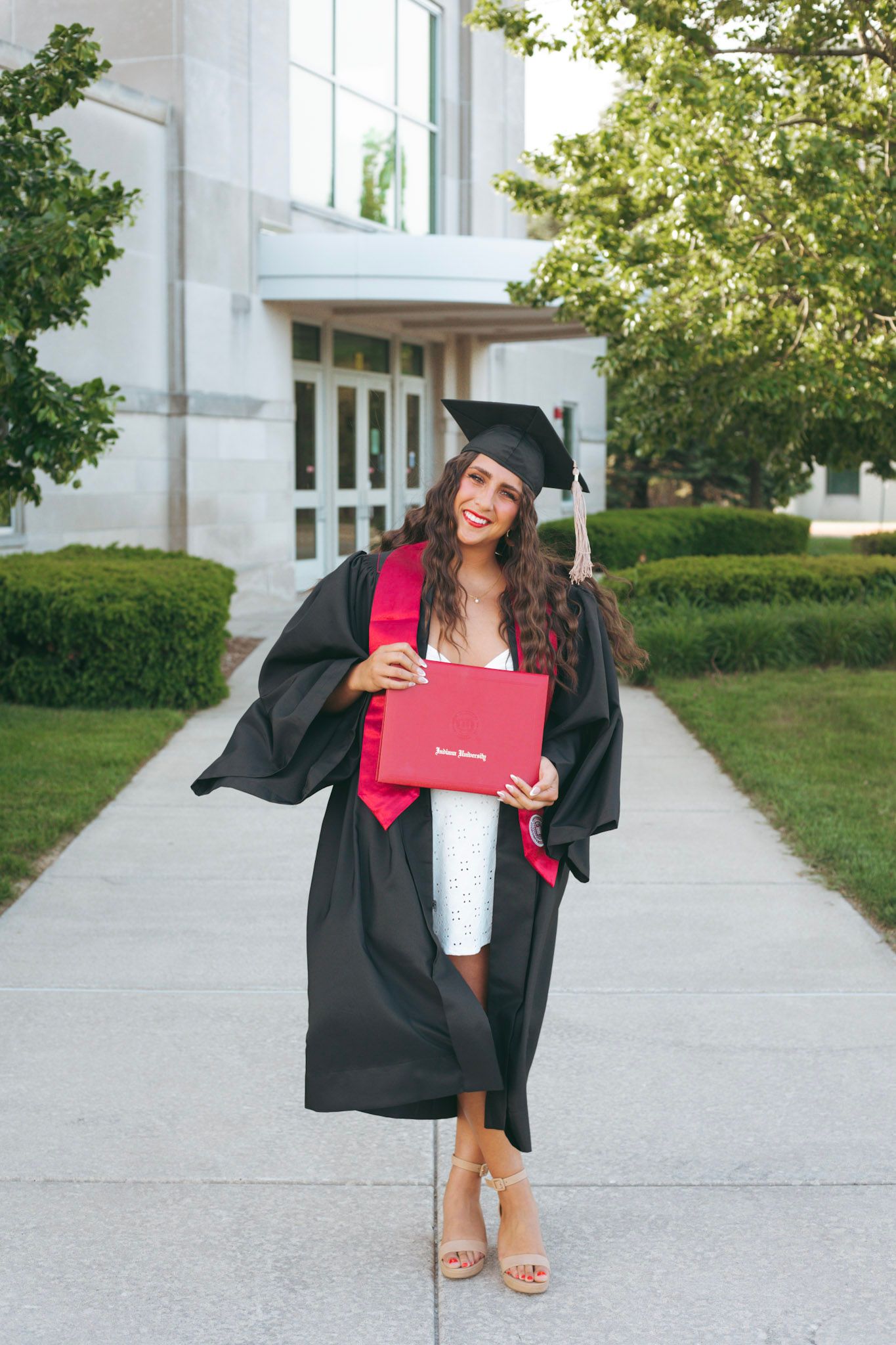 Smiling female graduate in black cap and gown holding red diploma cover outside campus building.