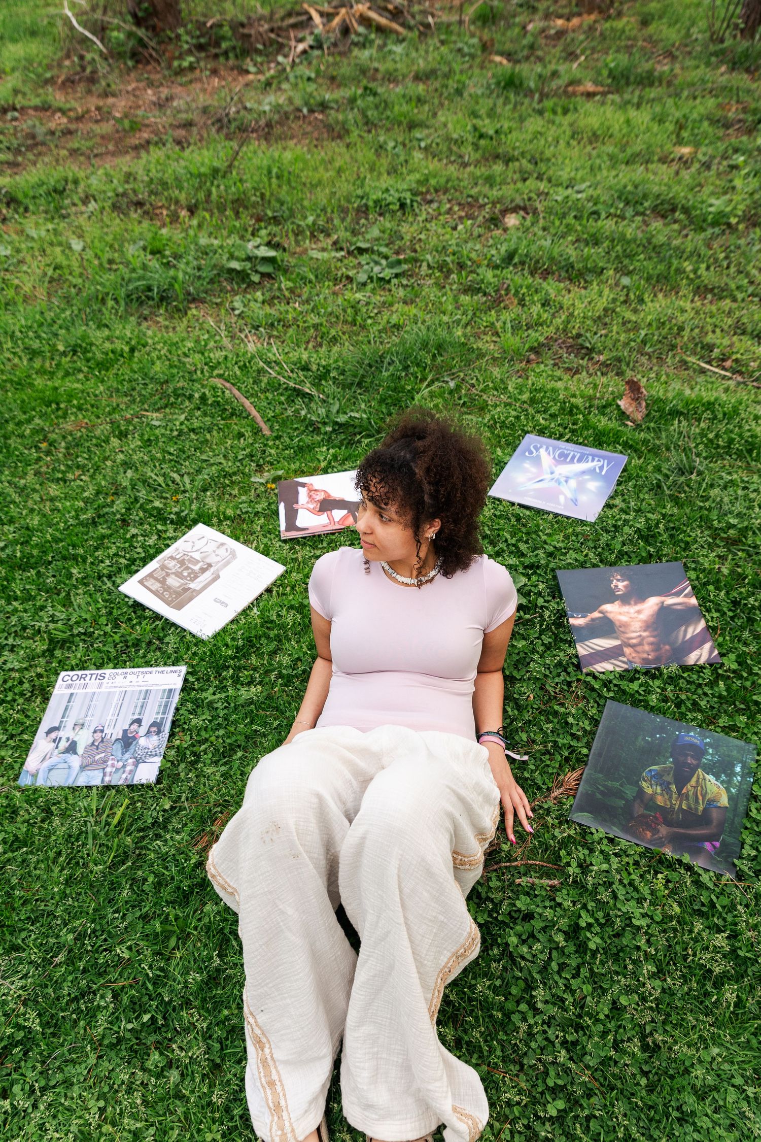 Young woman with curly hair sitting on grass surrounded by vinyl record albums in outdoor setting.