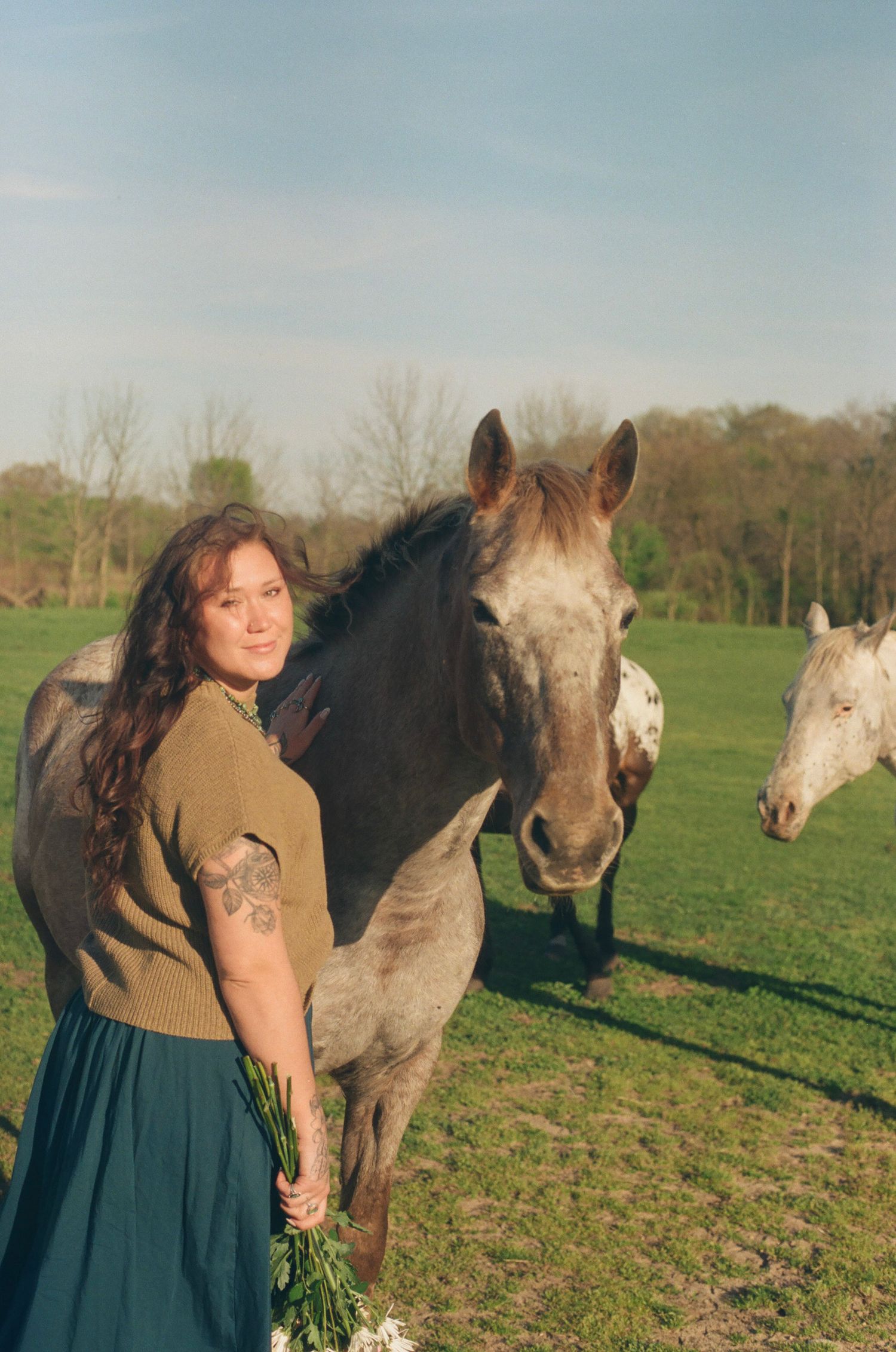 Woman with tattoos posing beside two gray horses in a sunny green farm field.