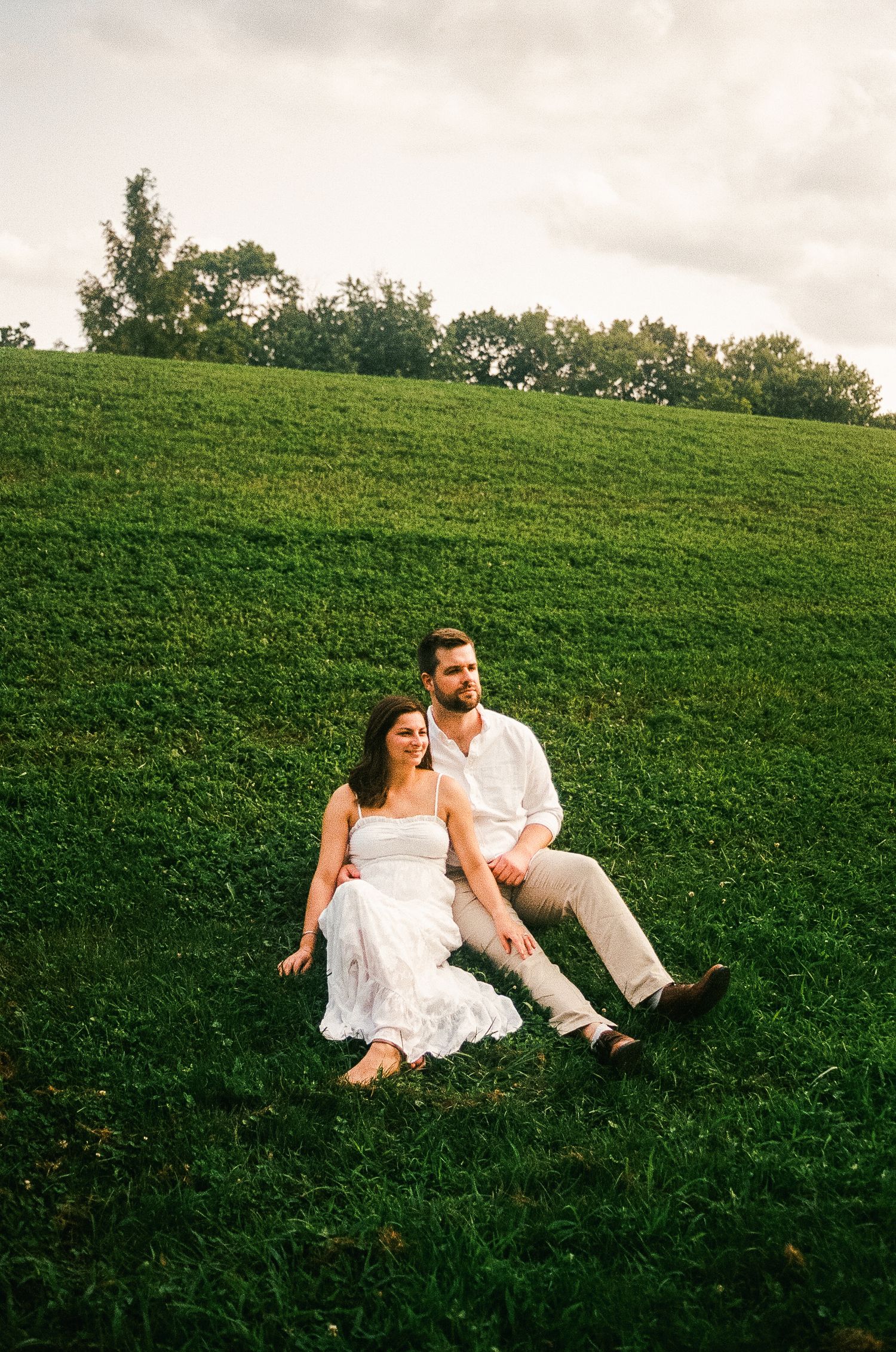 Couple in white outfits sitting on lush green hillside, woman in white dress leaning against man in white shirt.