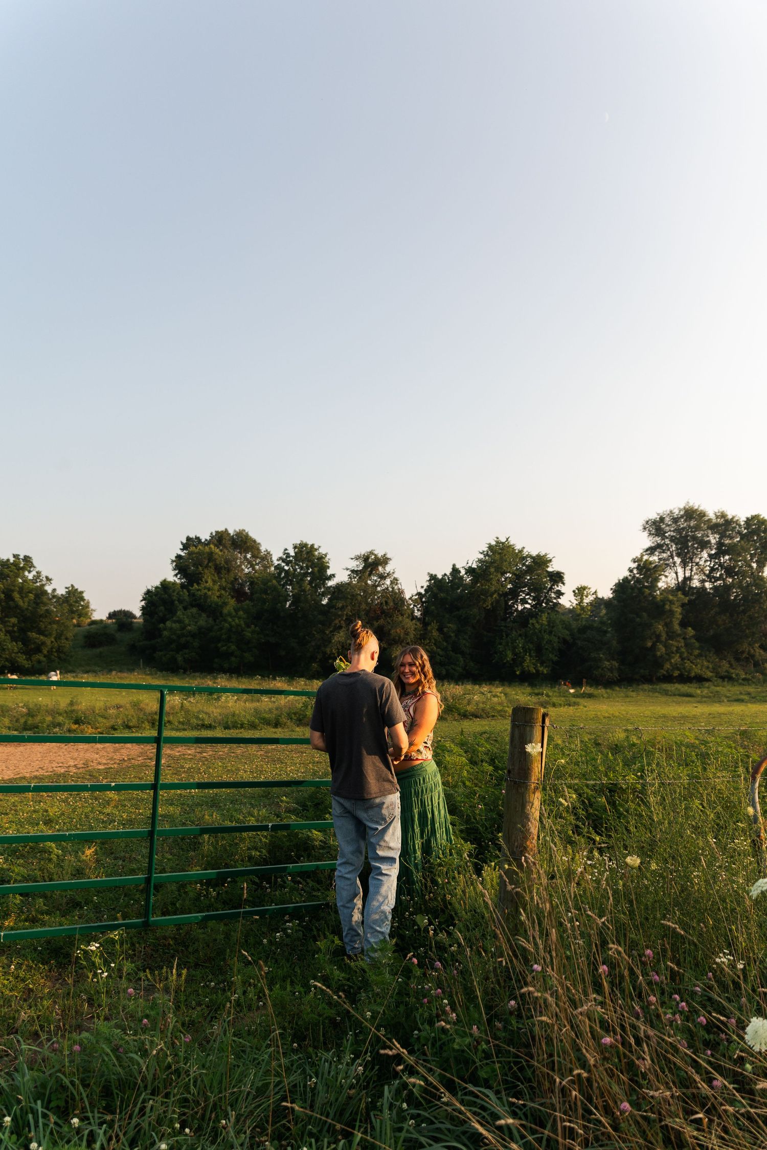Couple standing together by a green farm gate in a lush rural field at golden hour sunset.