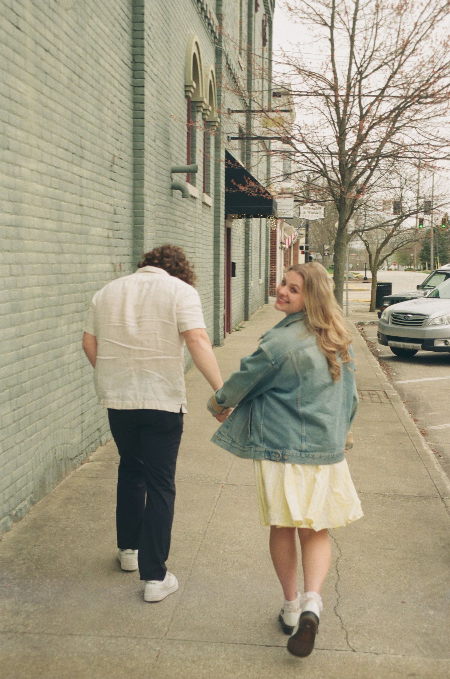 Woman in denim jacket smiles back at camera while holding hands with man on a brick sidewalk.