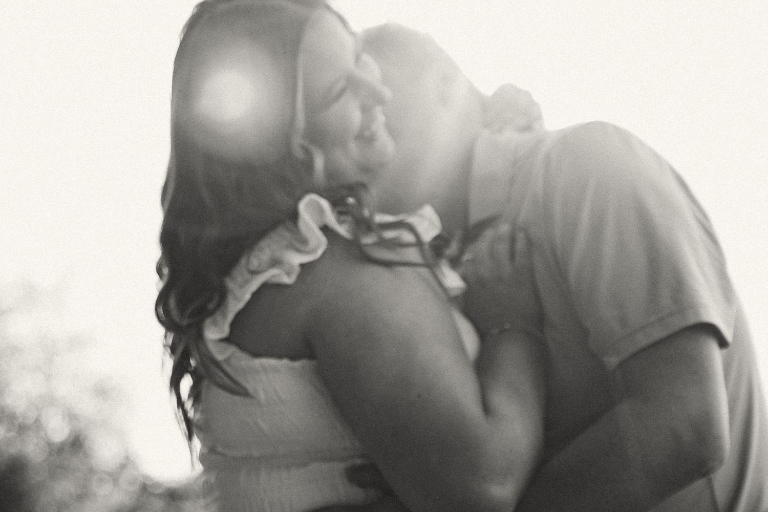 Romantic black and white photo of couple sharing a kiss in soft backlit sunlight outdoors.