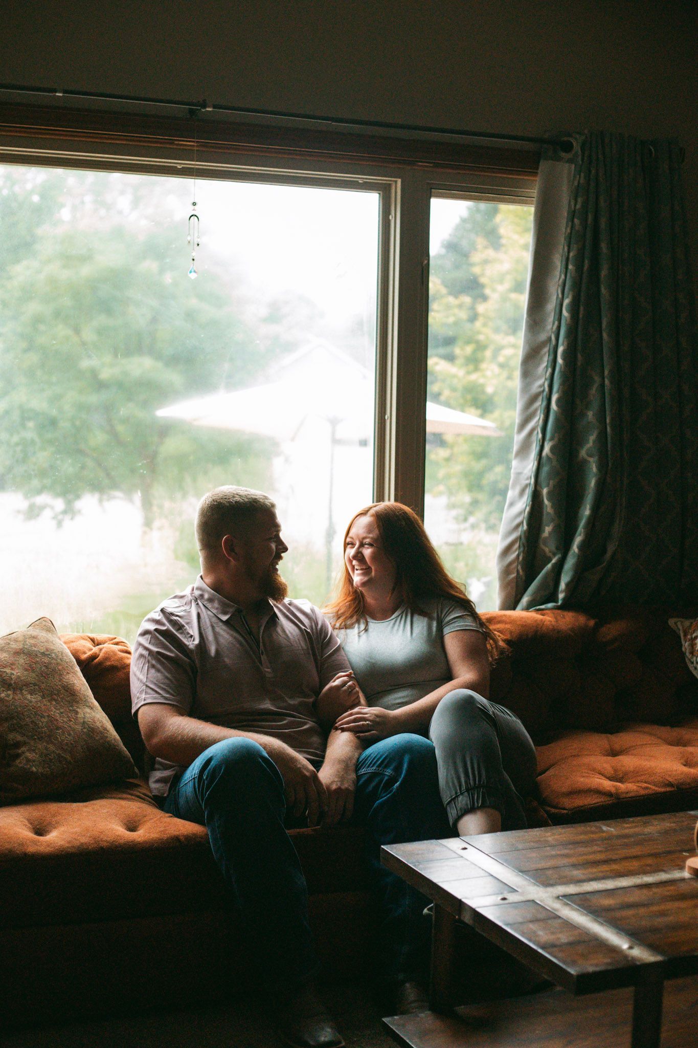 A happy couple sits on an orange couch laughing together in front of a large window with green trees outside.