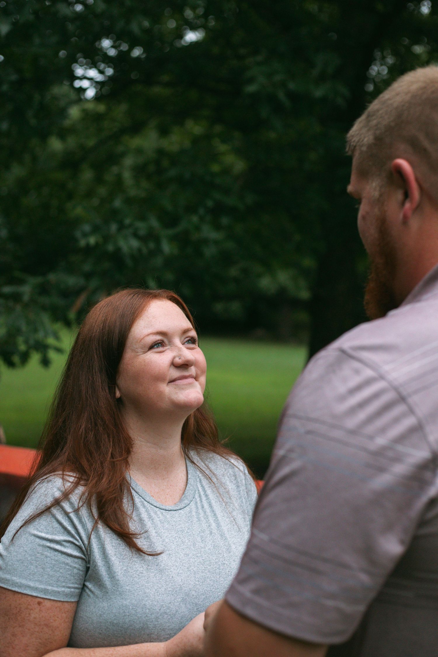 A smiling redheaded woman gazes up at her partner while standing outdoors with lush green trees in the background.