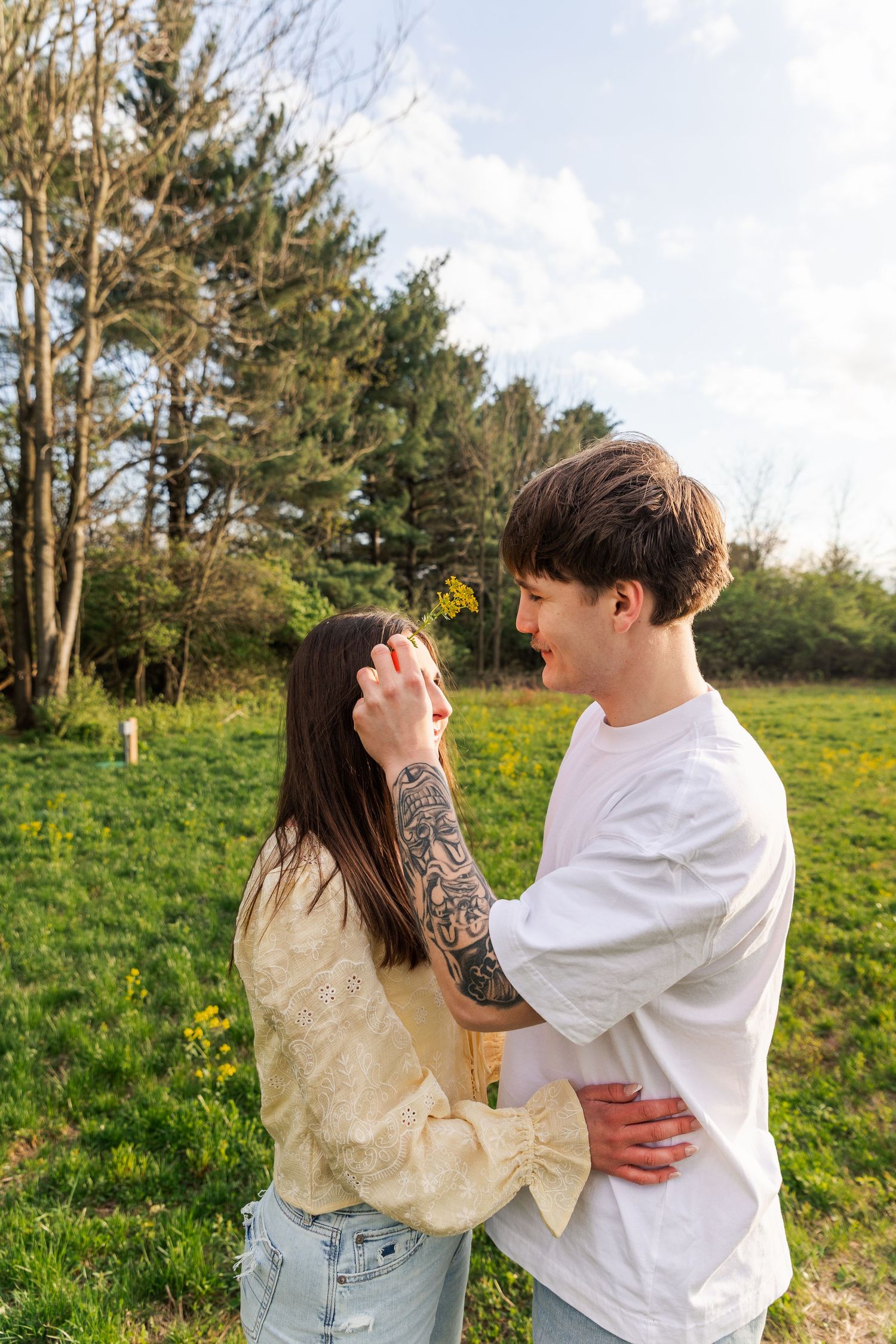 Tattooed man placing flower in woman's hair while embracing in a sunny green field.