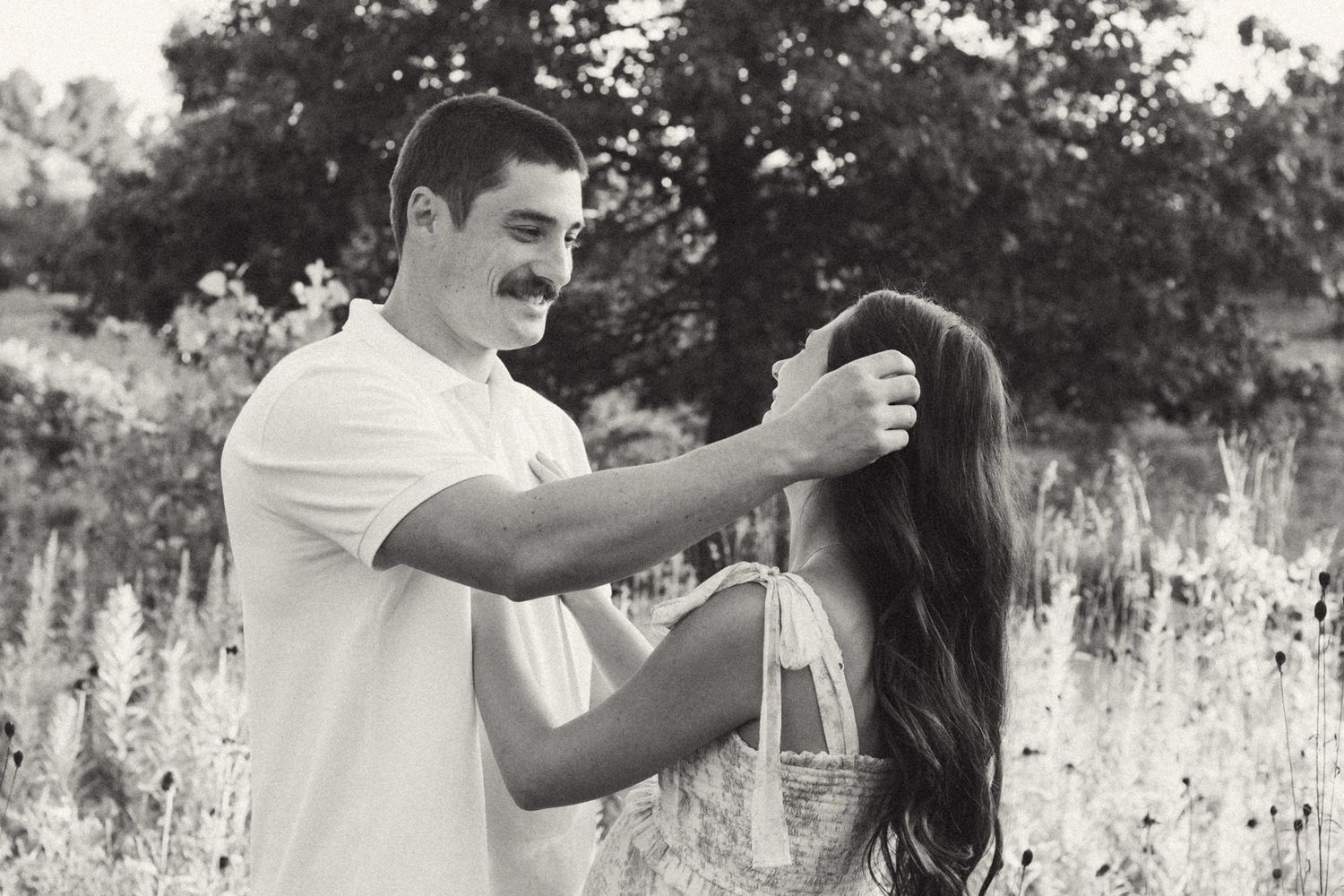 A black and white photo of a man gently placing flowers in a long-haired womans hair in a sunny field.