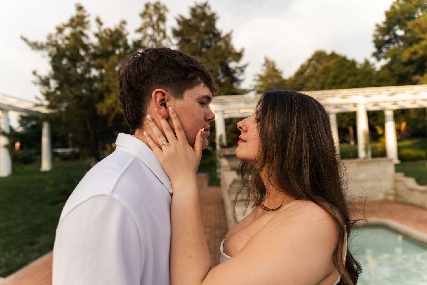Couple sharing a romantic moment near a pool, woman tenderly touching mans face, lush garden backdrop.