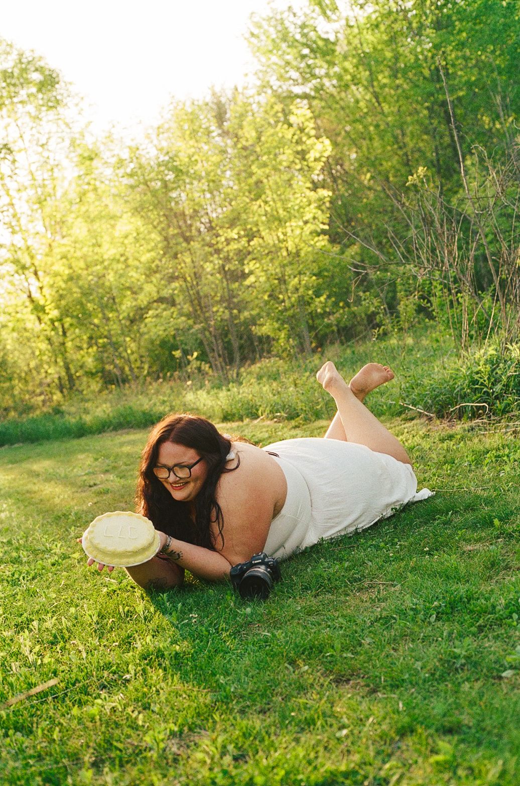 A smiling woman with glasses lies on green grass in a white dress, holding a straw hat outdoors at golden hour.