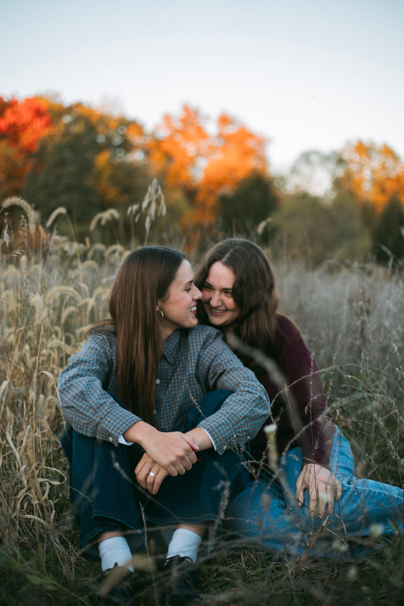 Two young women sitting close together in tall grass with colorful autumn trees behind.
