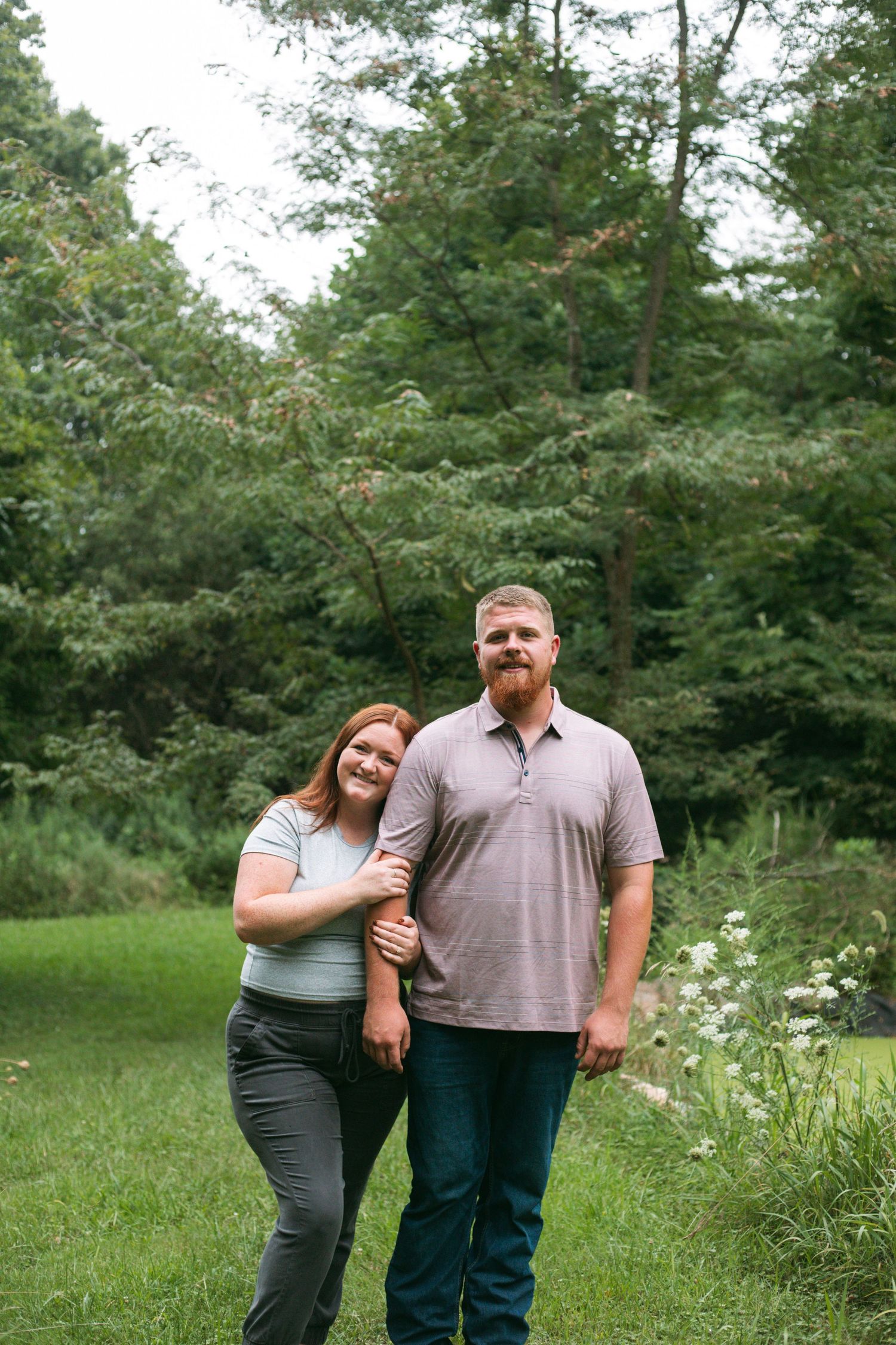 A smiling couple poses together in a lush green outdoor setting surrounded by tall trees and wildflowers.