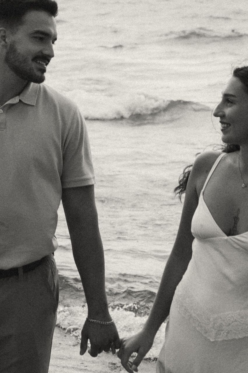 Black and white photo of a smiling couple holding hands on a beach with ocean waves in the background.